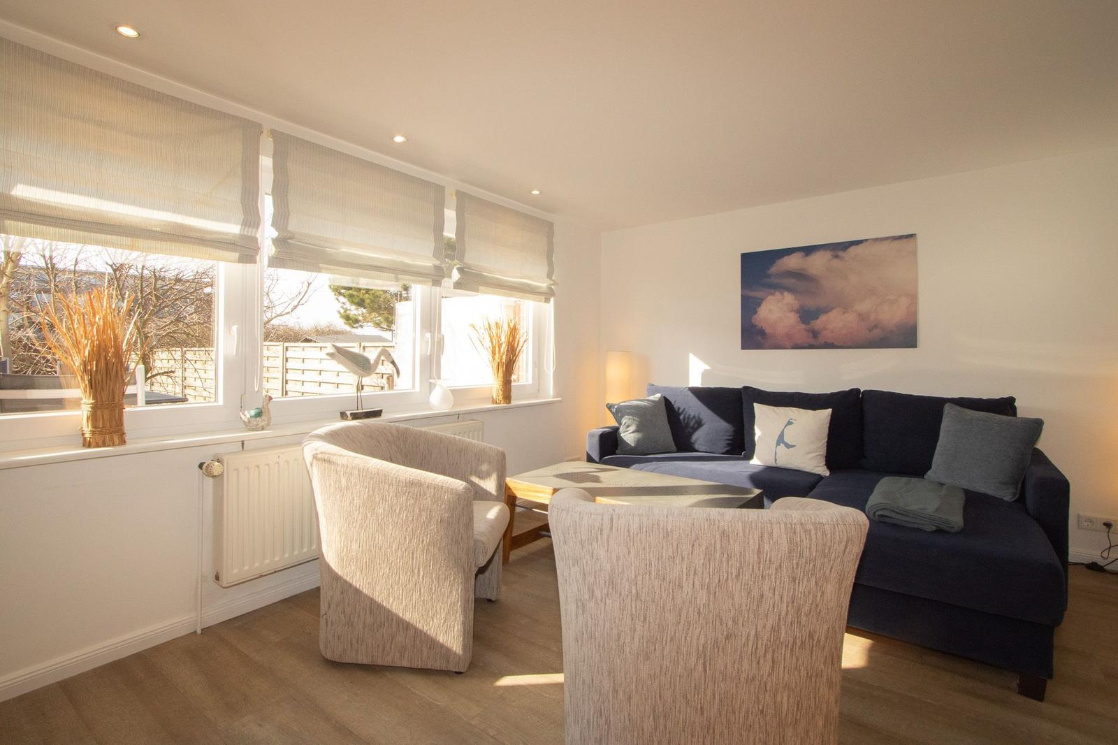 Wohnzimmer mit Sofa, zwei Sesseln und Fenstern mit Jalousien.