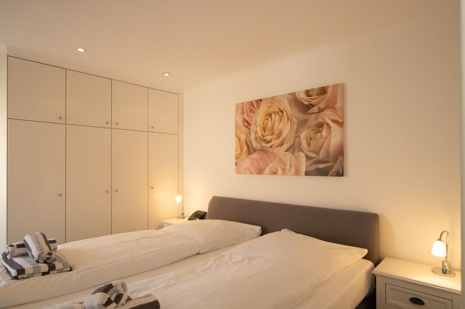 Schlafzimmer mit Doppelbett, Kleiderschrank und Blumenbild an der Wand.