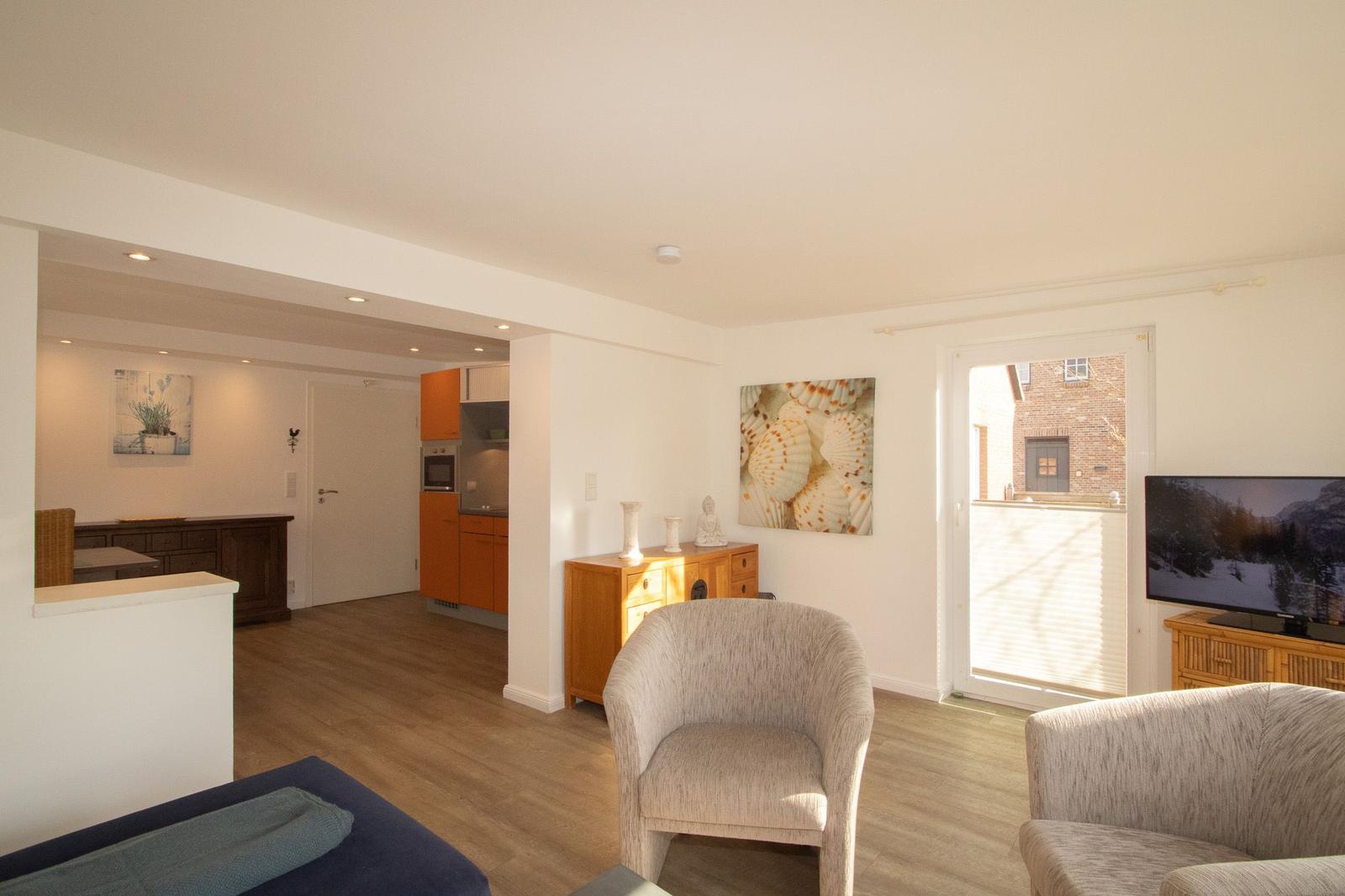 Wohnzimmer mit Sitzgelegenheiten, Fernseher und Blick auf Balkon. Küche im Hintergrund.