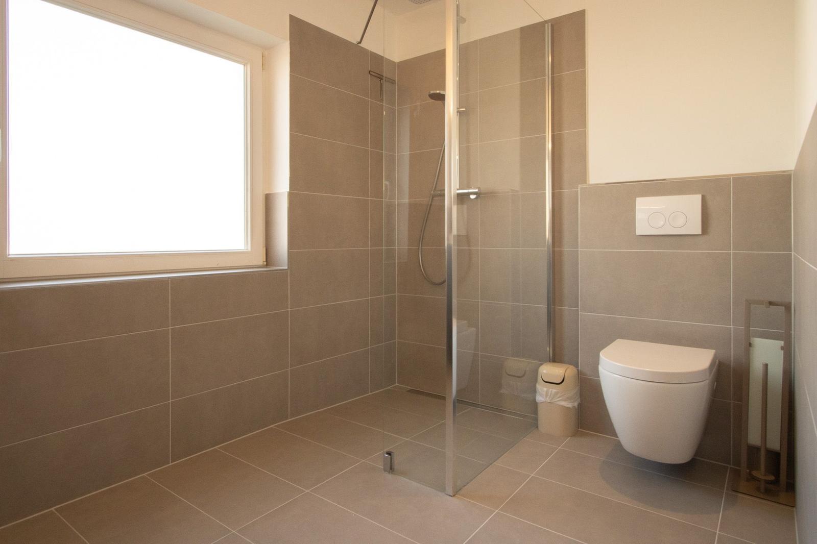 Badezimmer mit Dusche, WC und Fenster. Wand- und Bodenfliesen grau.
