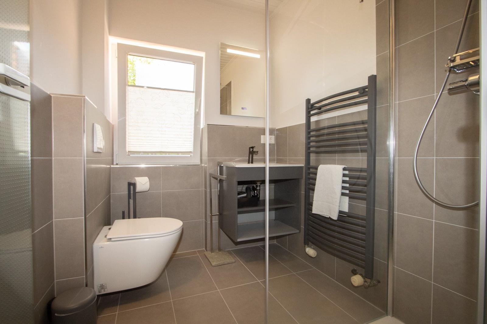 Bathroom with shower, toilet, and sink. Window with blinds and towel rack.