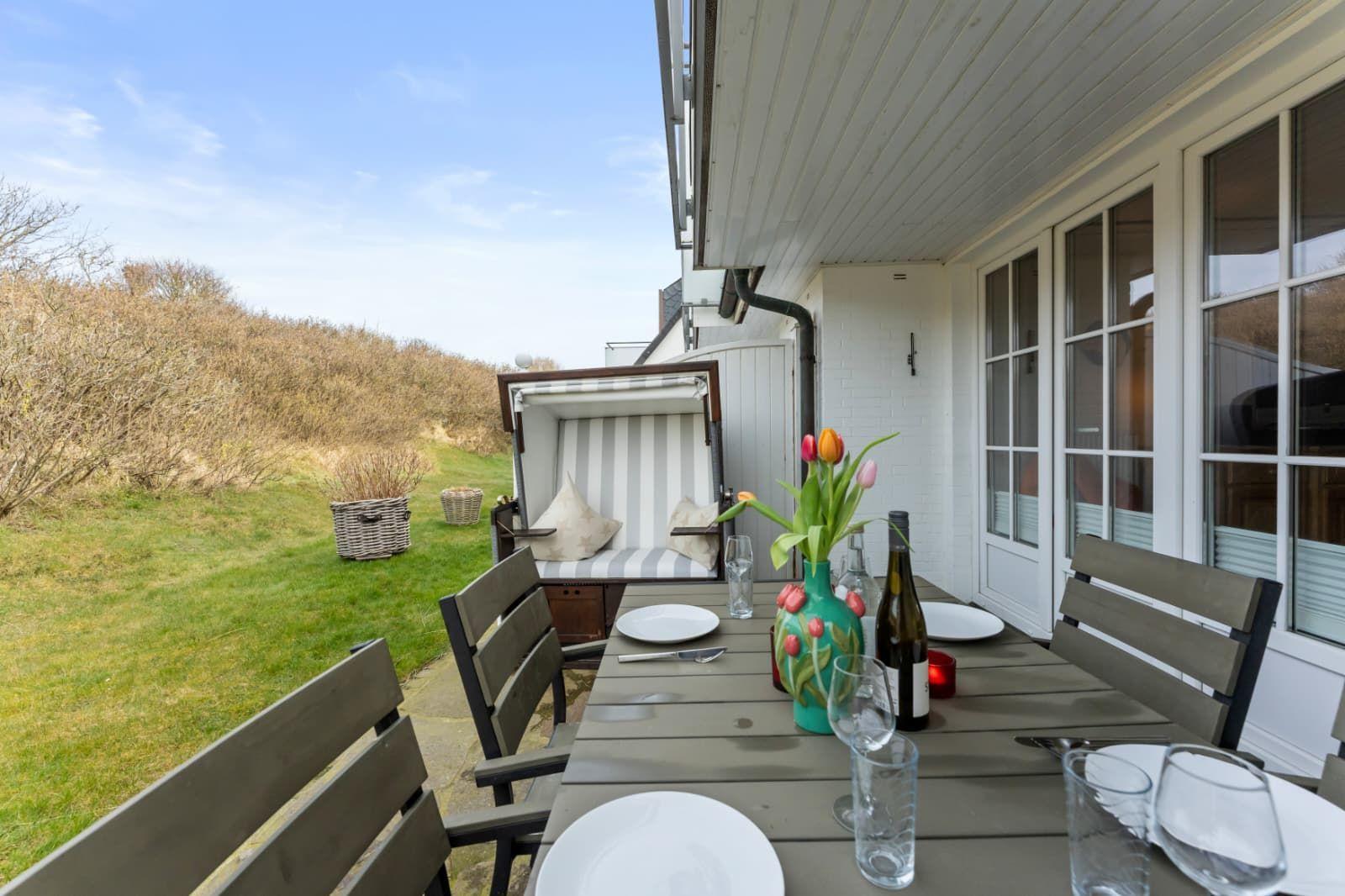 Terrasse mit Tisch, Stühlen und Blumen. Hintergrund: Grasfläche und Hecke.