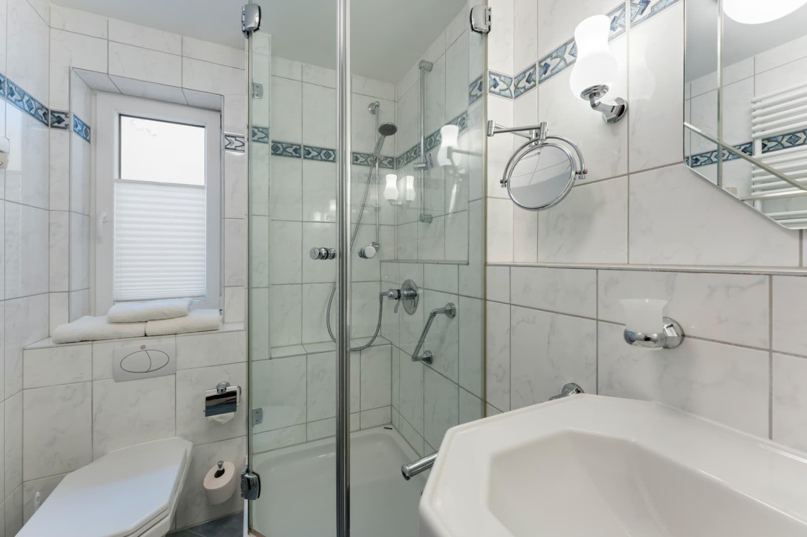 Badezimmer mit Dusche, Waschbecken und Toilette. Weiße Fliesen und Fenster mit Jalousie.
