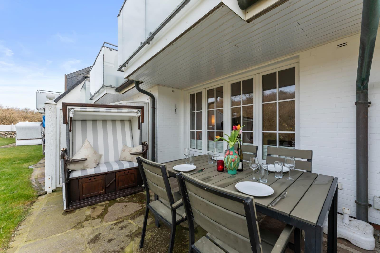 Terrasse mit Esstisch, Stühlen und Strandkorb. Hintergrund: weißes Haus mit Fenstern.