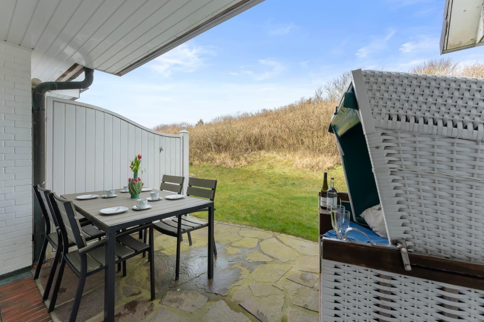 Terrace with table, chairs, and beach chair. Background: grassy area and hedge.