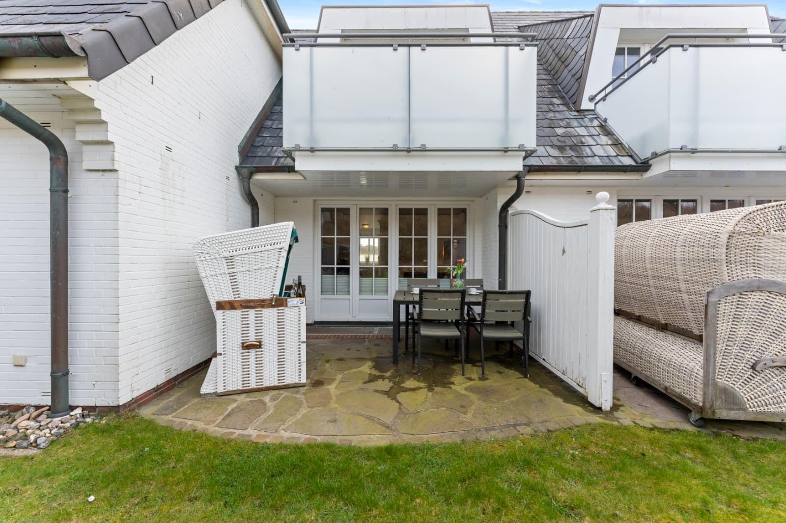 Backyard with table, chairs, wicker furniture, and garden gate. White wall and stone paving.