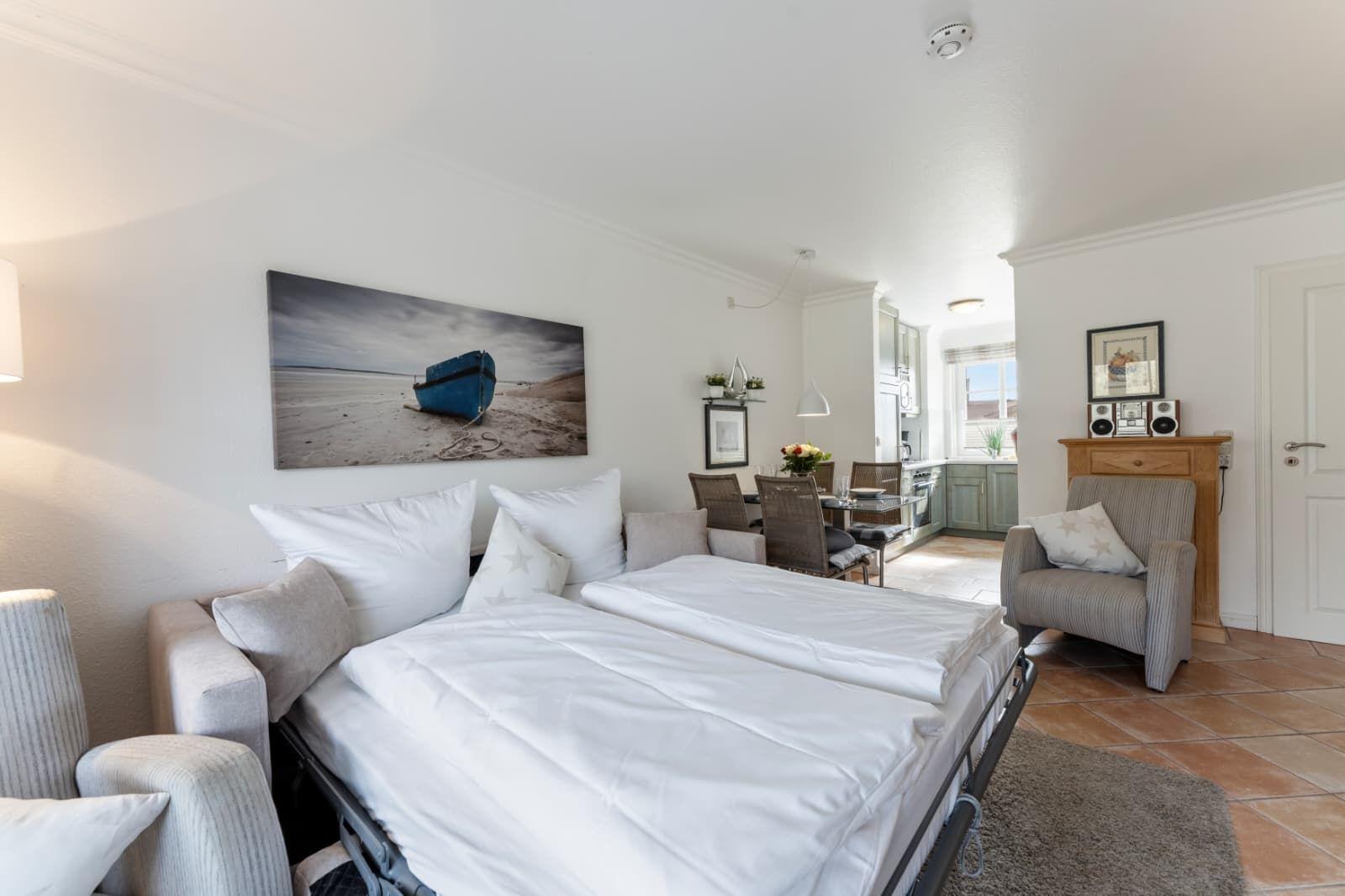 Bedroom with double bed, armchairs, and wall art. Kitchen and dining area are visible.