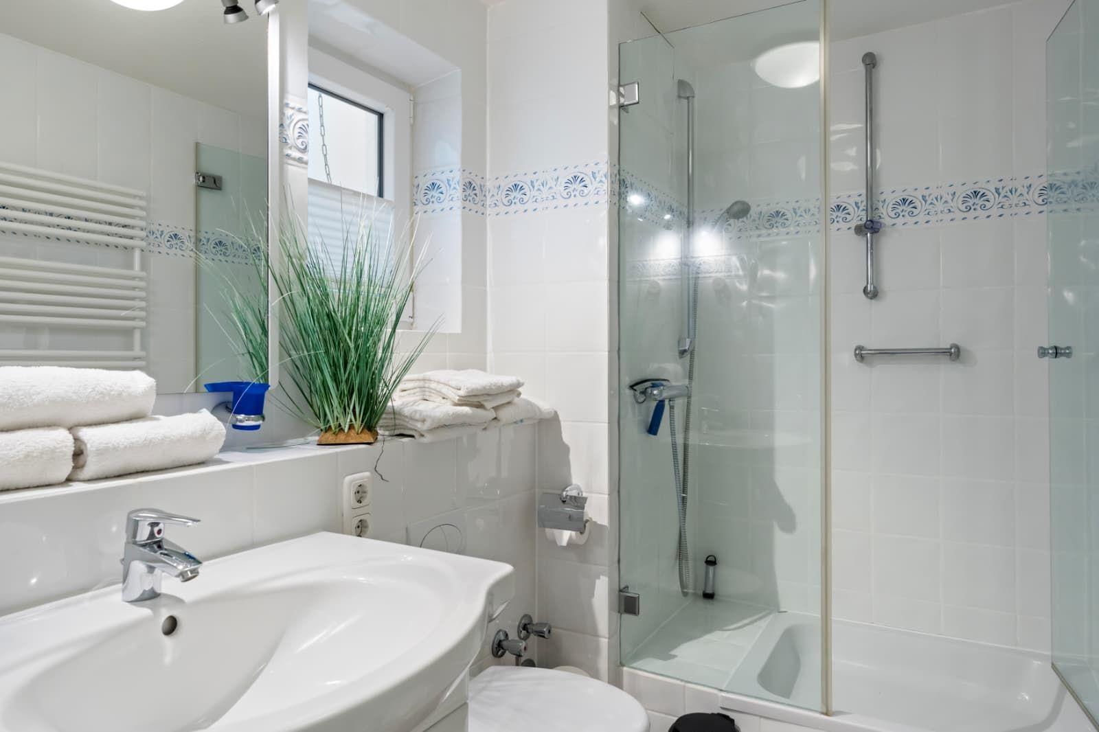 Bathroom with shower, sink, and toilet. White tiles and plant visible.