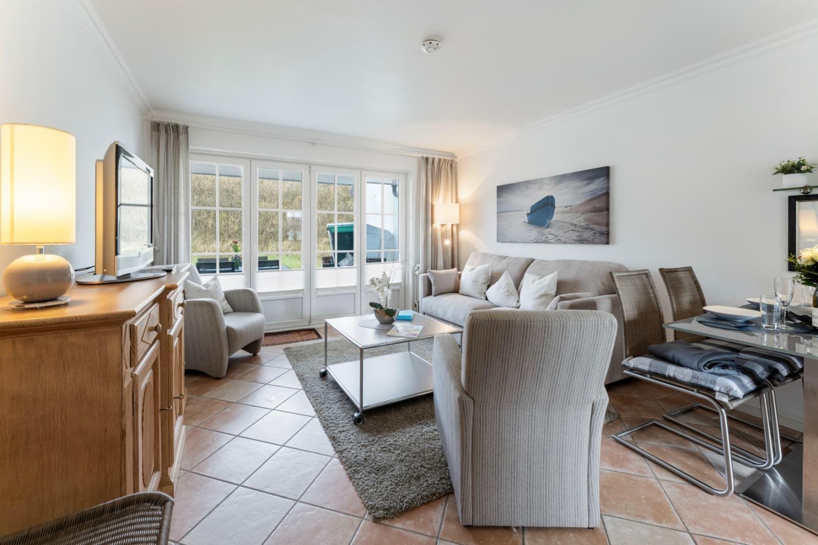 Living room with sofa, armchairs, dining table, and TV. Large window front with outdoor view.