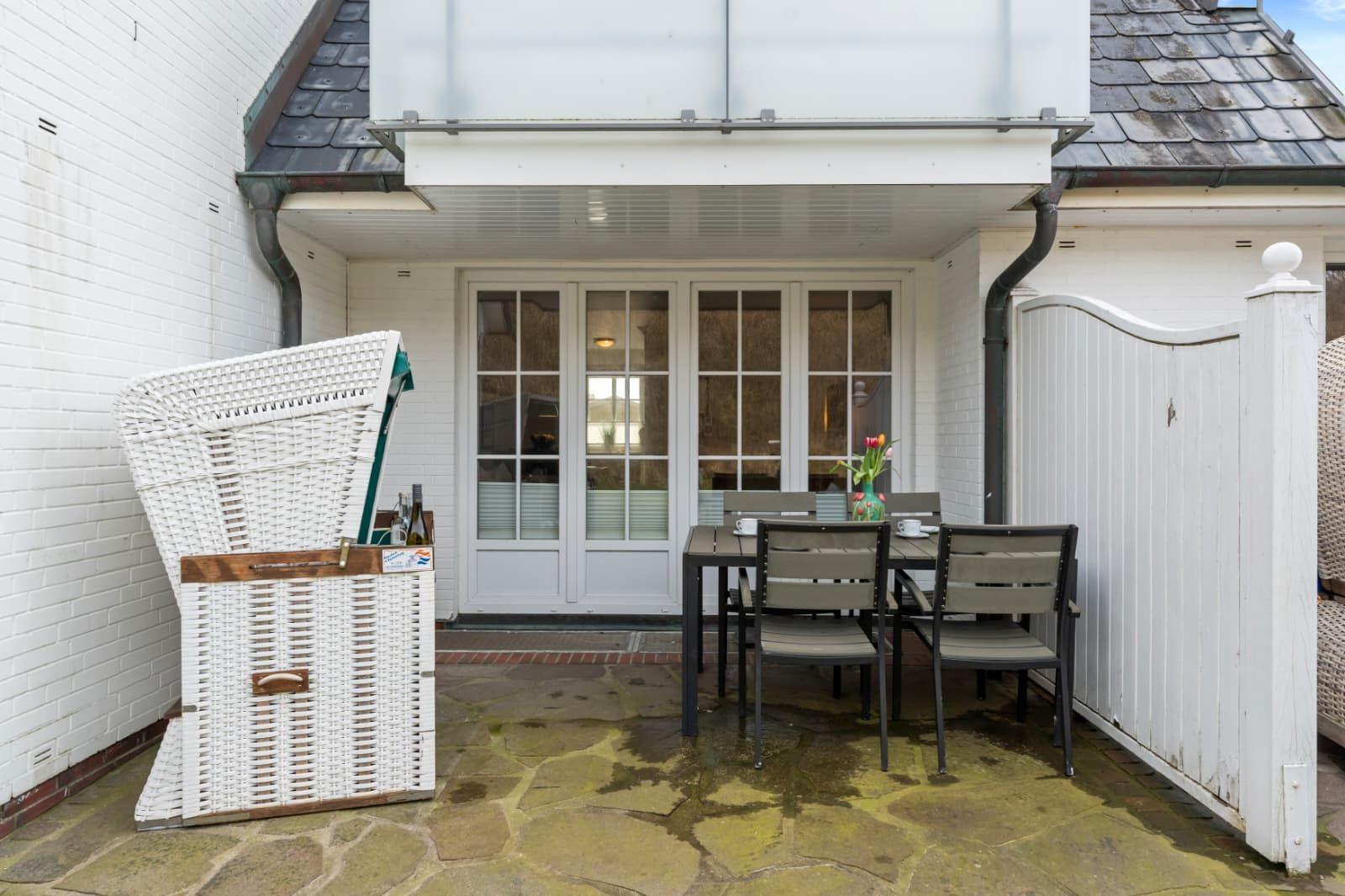 Terrace with table, chairs, and white beach chair. Background: white house wall and glass doors.