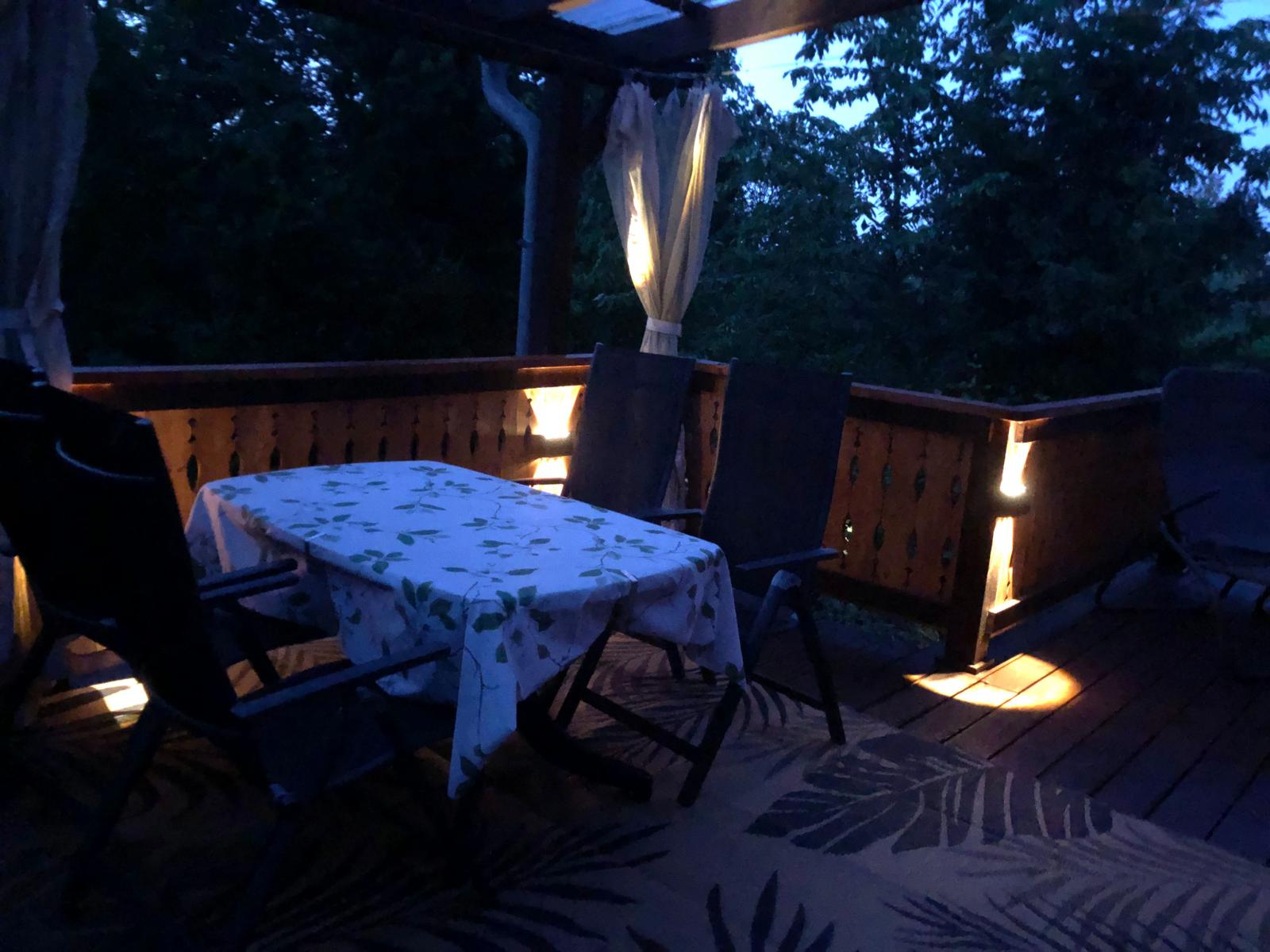 Terrasse mit Tisch, Stühlen und Beleuchtung. Hintergrund: dunkler Wald.
