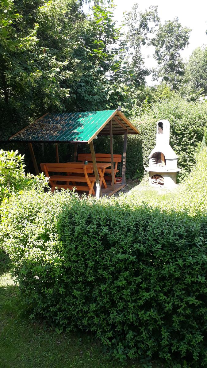 Garten mit Holzüberdachung und Grill. Umgeben von grünen Sträuchern und Bäumen.