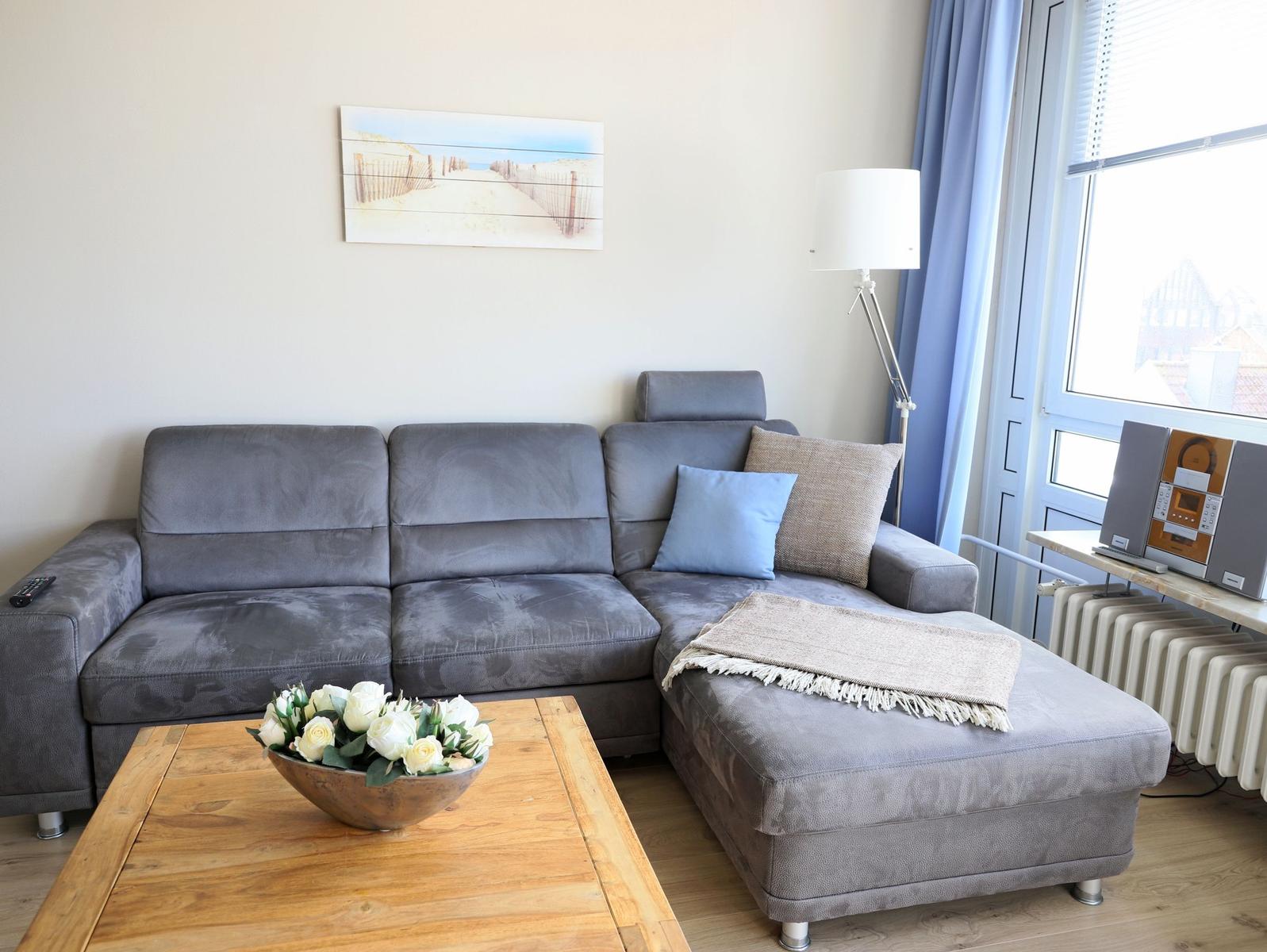 Ein grauer Sofa mit Kissen und Decke. Holztisch mit Blumen. Fenster mit Vorhängen.
