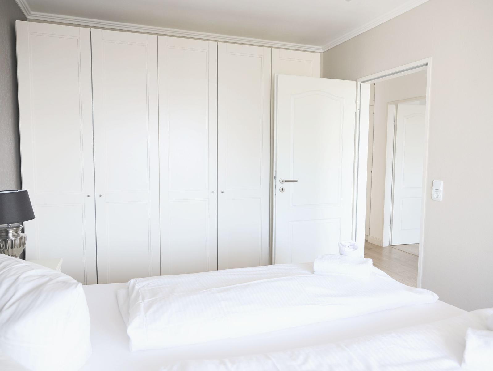 Schlafzimmer mit weißem Bett, Kleiderschrank und Tür zum Flur.