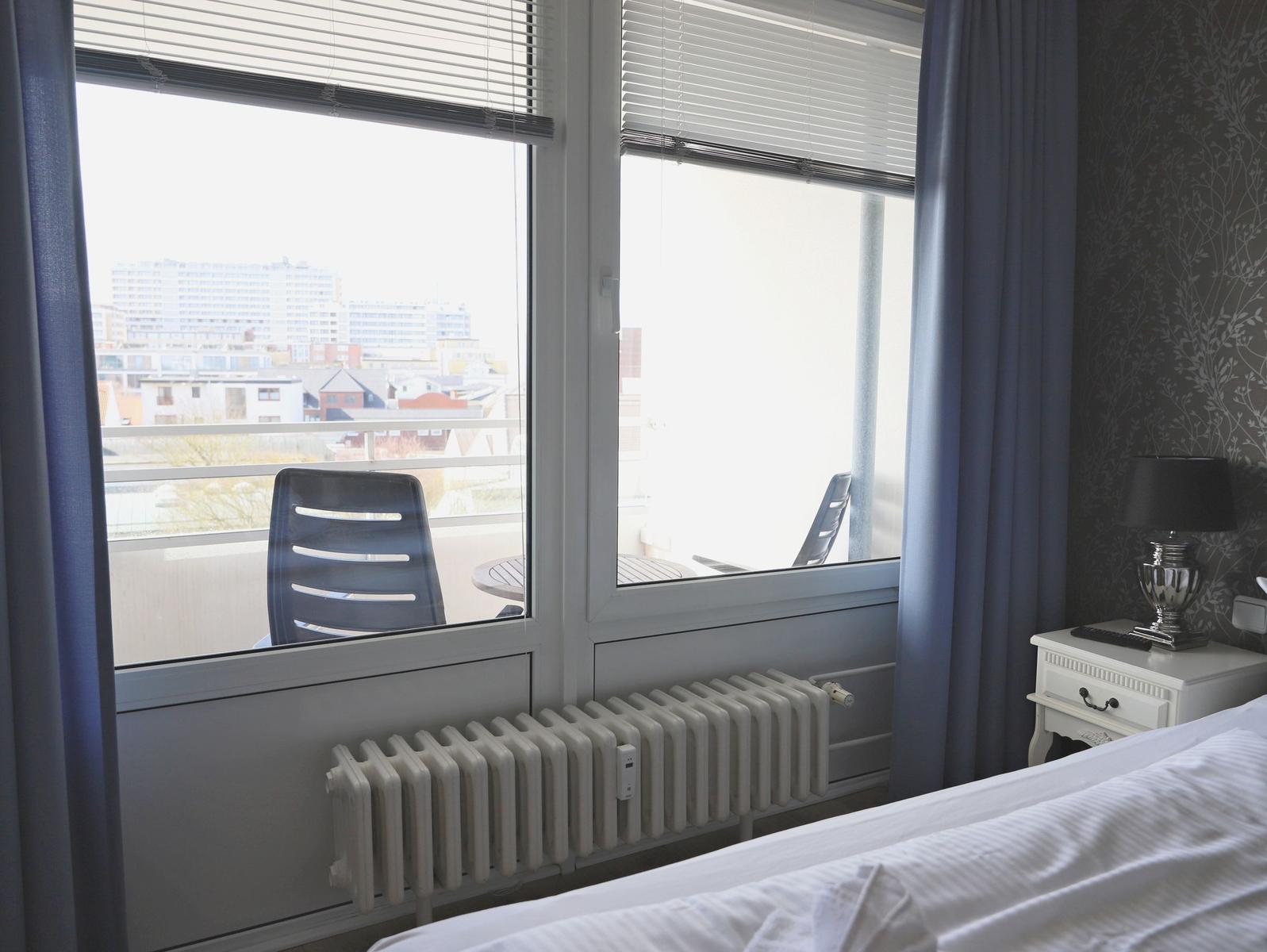 Zimmer mit Blick auf Balkon und Stadt. Bett, Nachttisch und Heizung sichtbar.