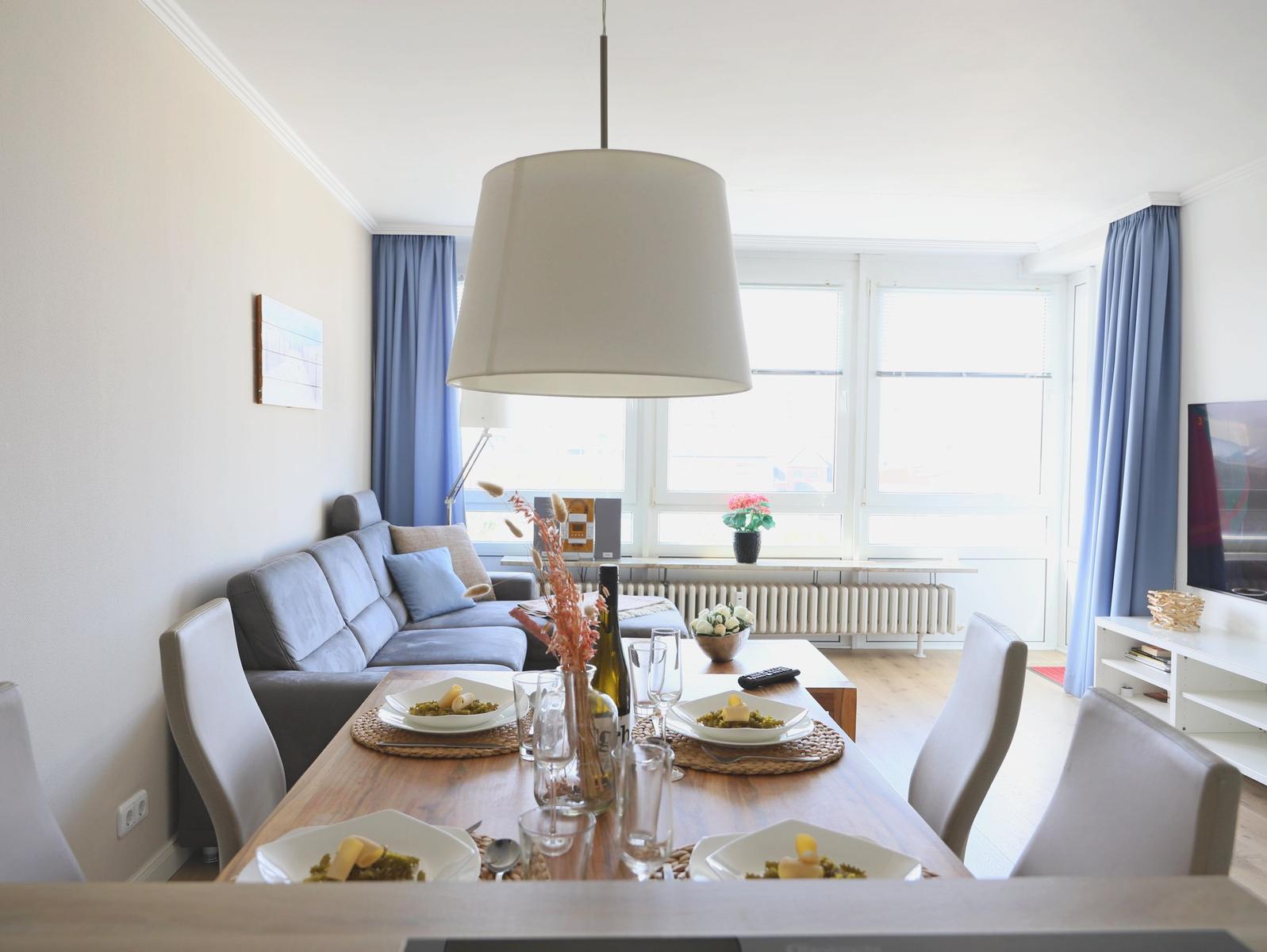 Essbereich mit Tisch, Stühlen und Sofa. Fenster mit blauen Vorhängen. Deckenleuchte über Tisch.