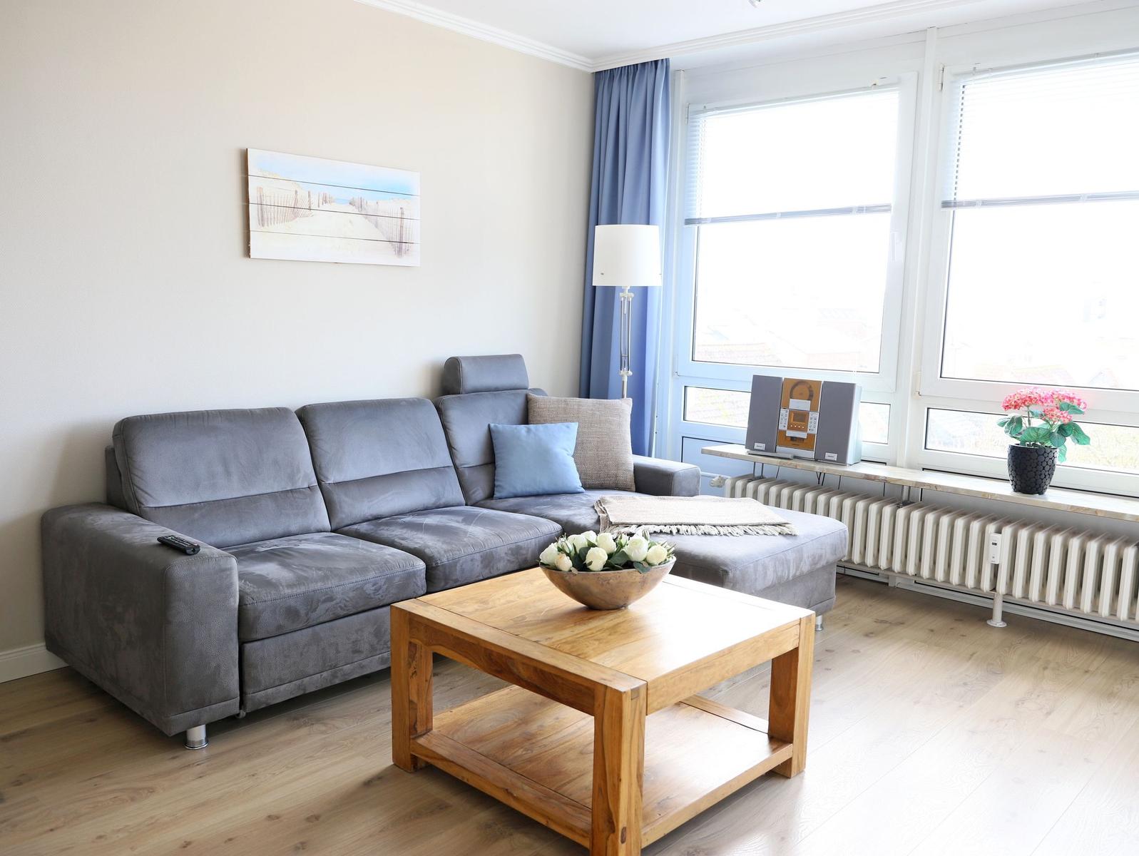 Ein Wohnzimmer mit grauem Sofa, Holztisch und Fenstern mit blauen Vorhängen.