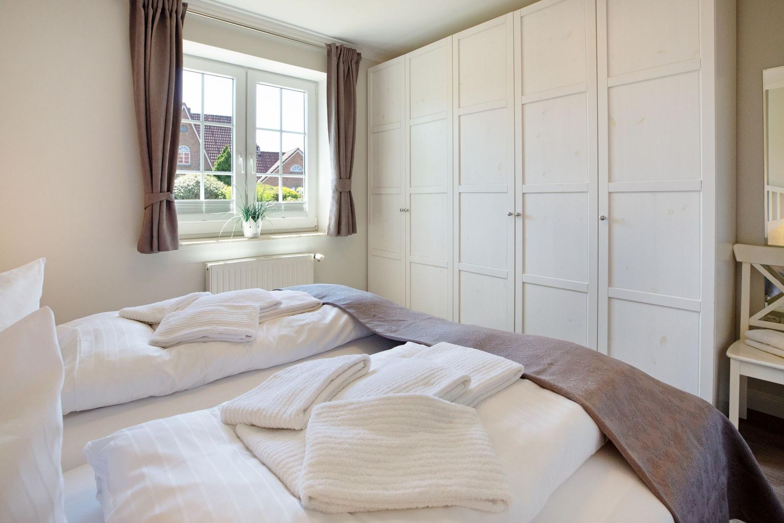 Schlafzimmer mit Doppelbett, weißen Wänden und großem Schrank. Fenster mit Vorhängen und Pflanze.