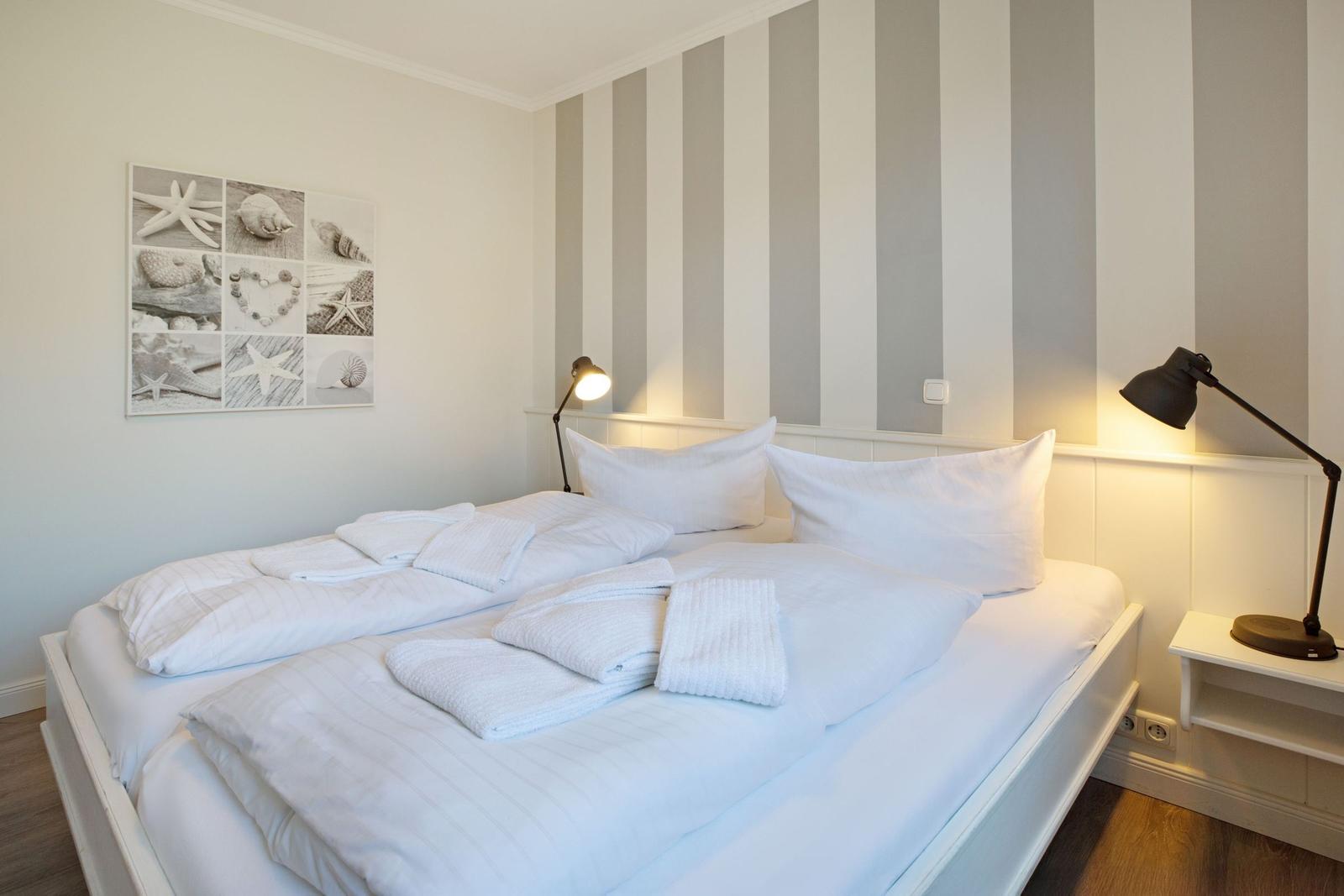 Schlafzimmer mit weißem Bett, Streifenwand und Strandmotivbild.
