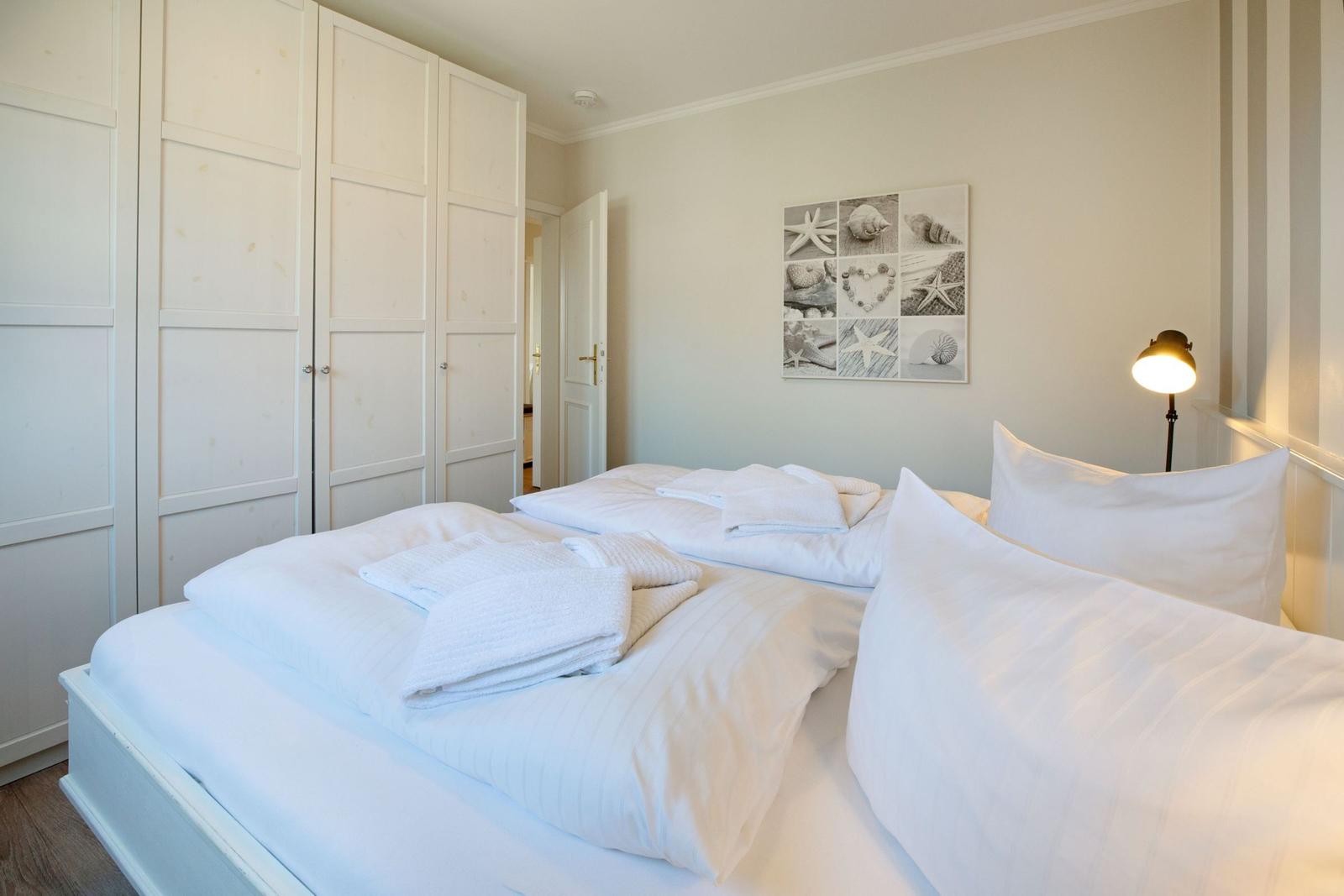 Schlafzimmer mit weißem Bett, Kleiderschrank und Wandbild mit Muscheln.