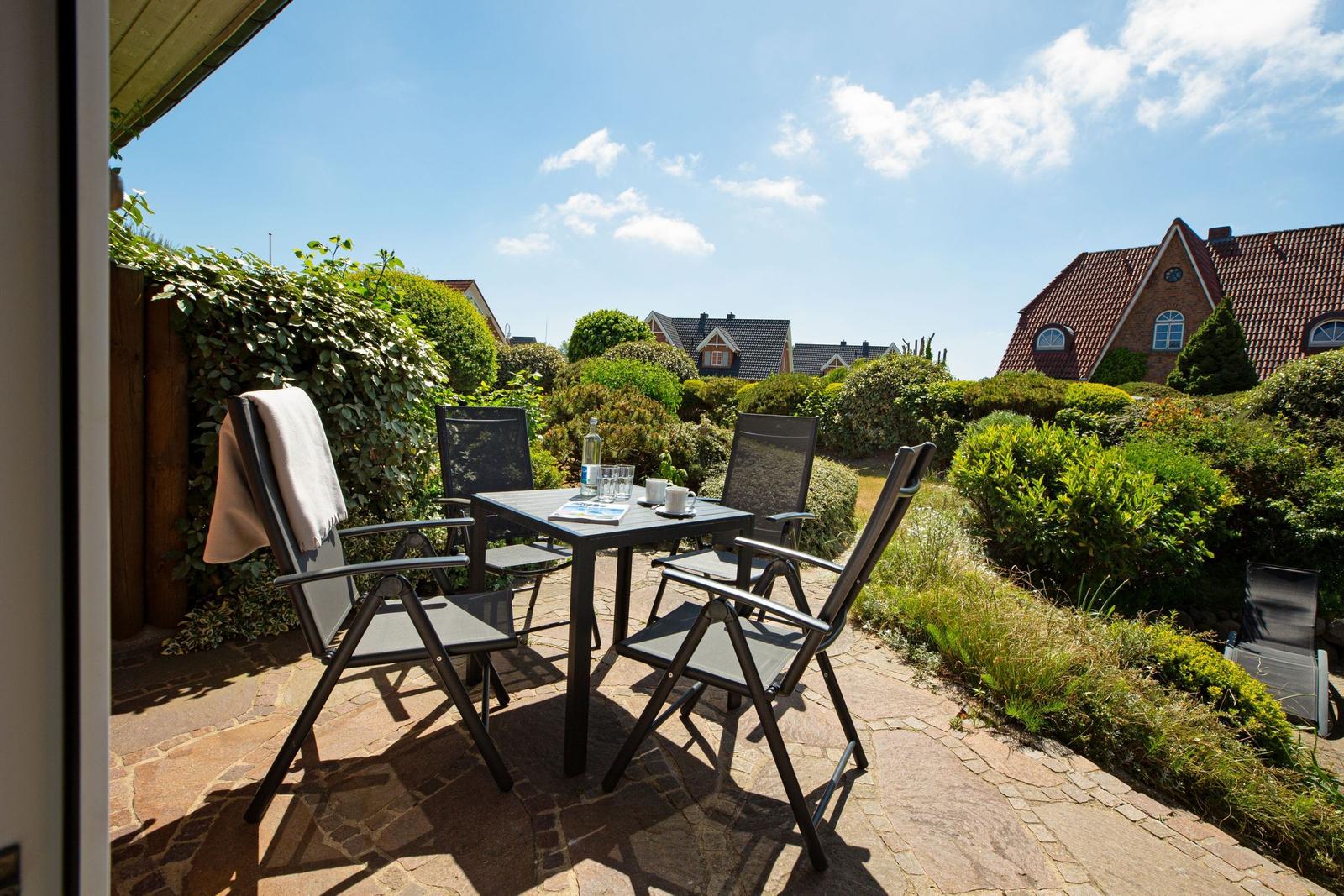 Terrasse mit Tisch und Stühlen, umgeben von grünen Sträuchern und Häusern im Hintergrund.