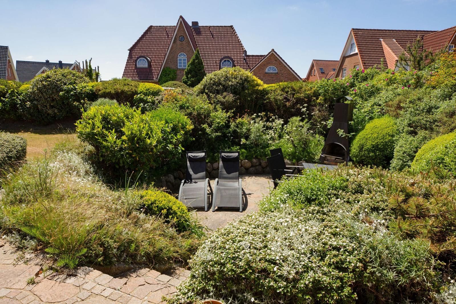Garten mit Liegestühlen und Grill, umgeben von grünen Sträuchern und einem gepflasterten Weg.