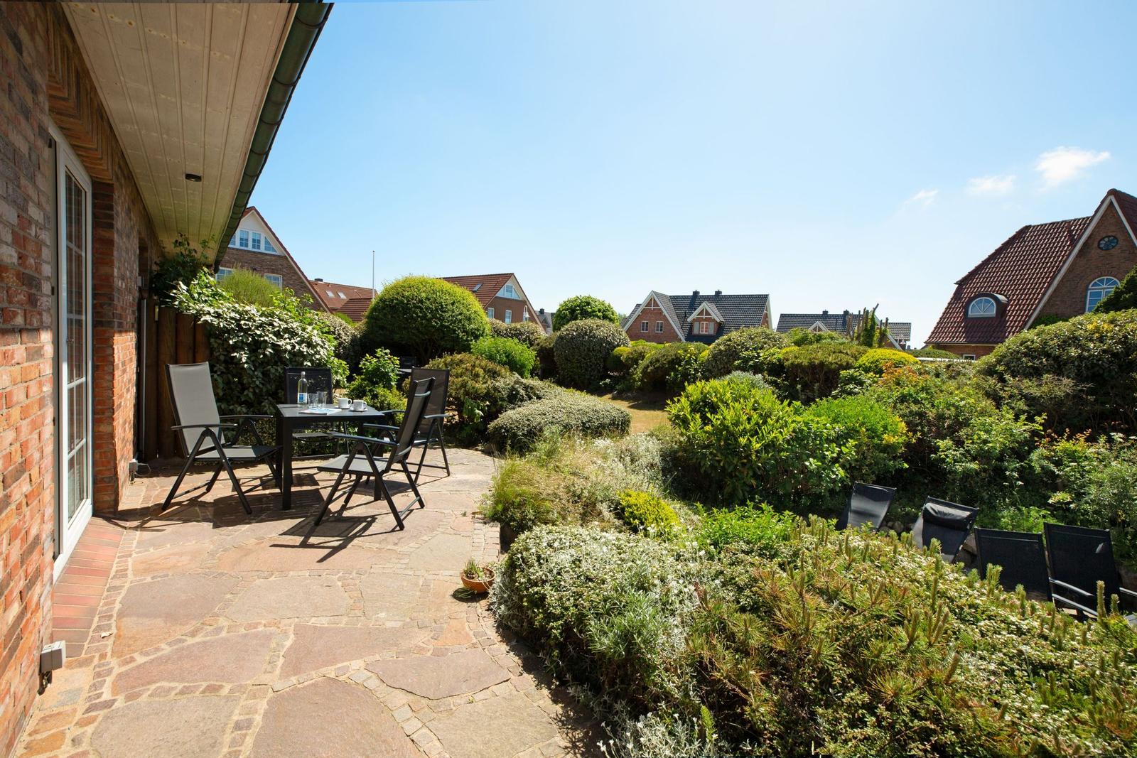 Terrasse mit Tisch und Stühlen, umgeben von grünen Sträuchern und einem gepflasterten Weg.