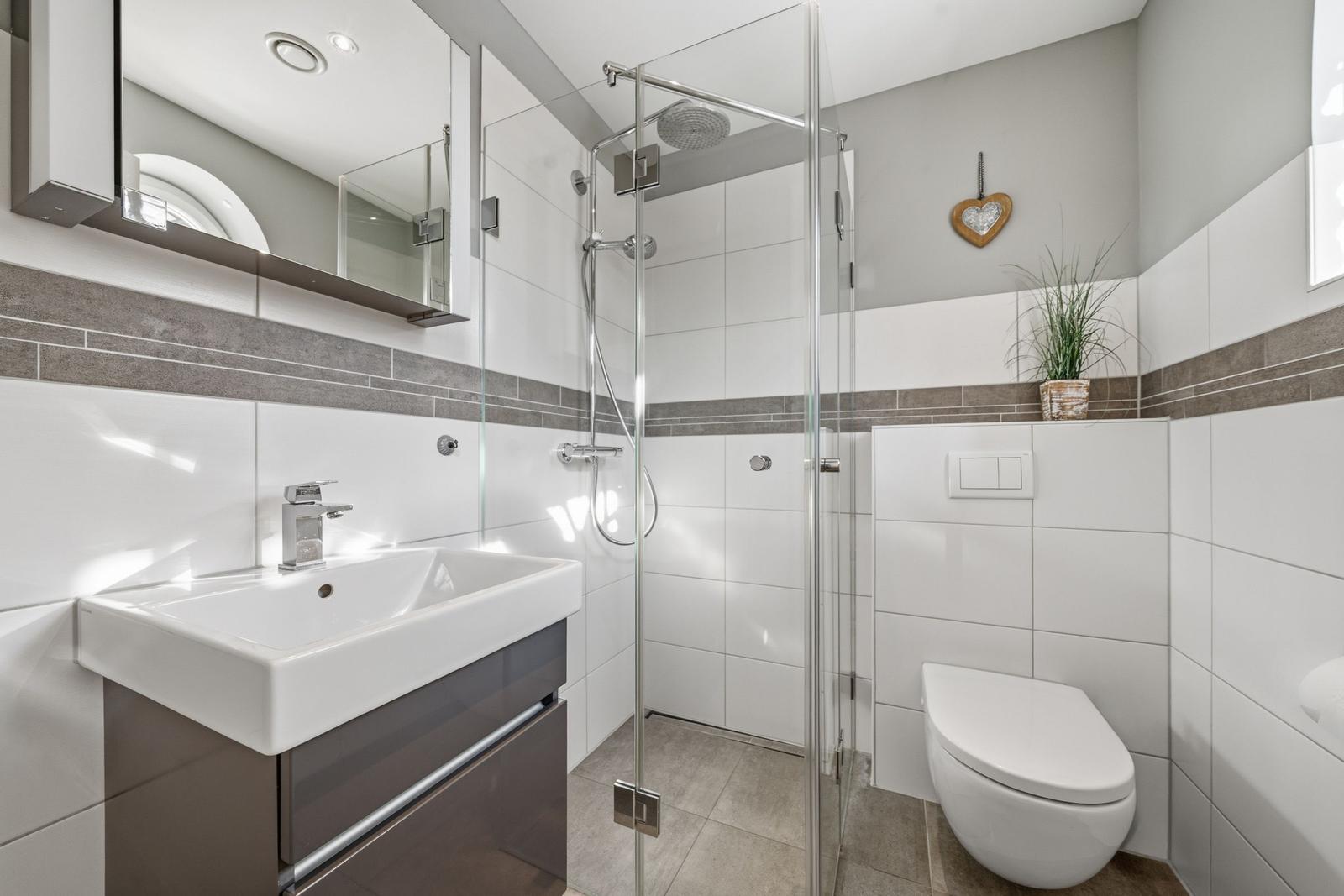 Badezimmer mit Dusche, Waschbecken und Toilette. Weiße Fliesen, Glasdusche und Pflanze.