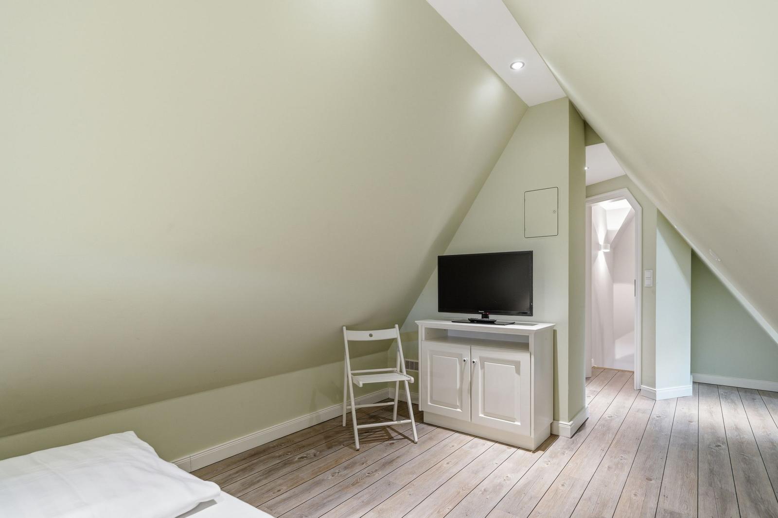 Zimmer mit Dachschrägen, weißer Kommode, Fernseher und Stuhl auf Holzboden.