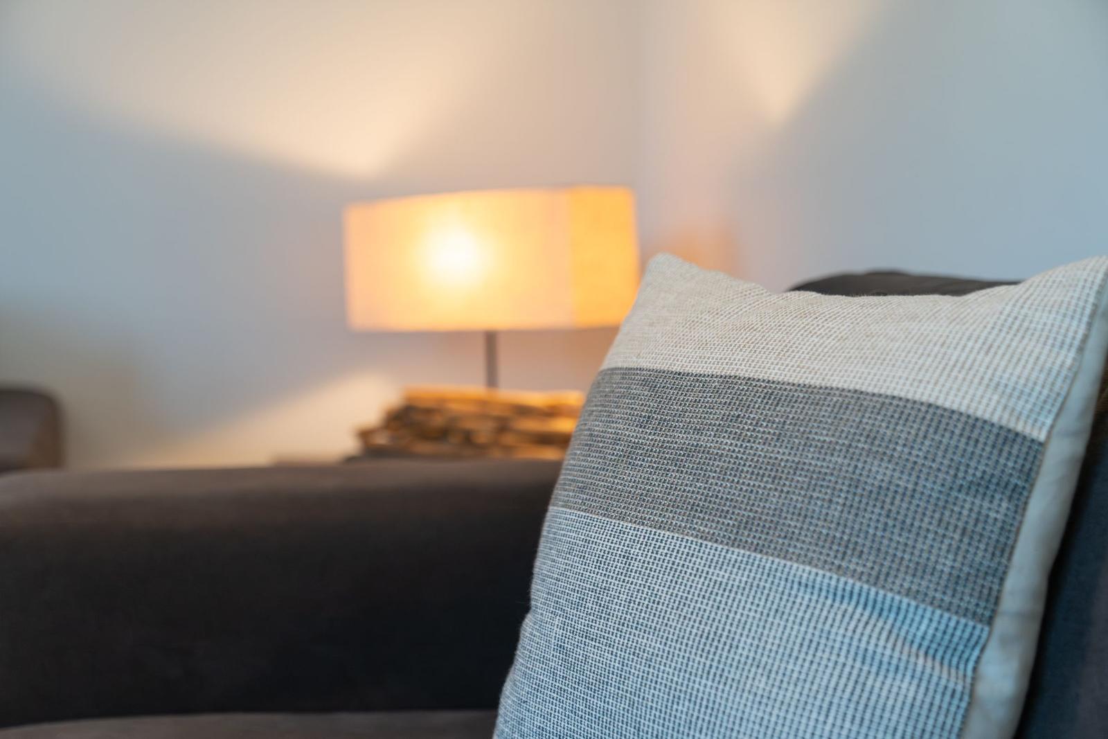 Ein Kissen mit Streifenmuster auf einem Sofa, dahinter eine Lampe mit warmem Licht.