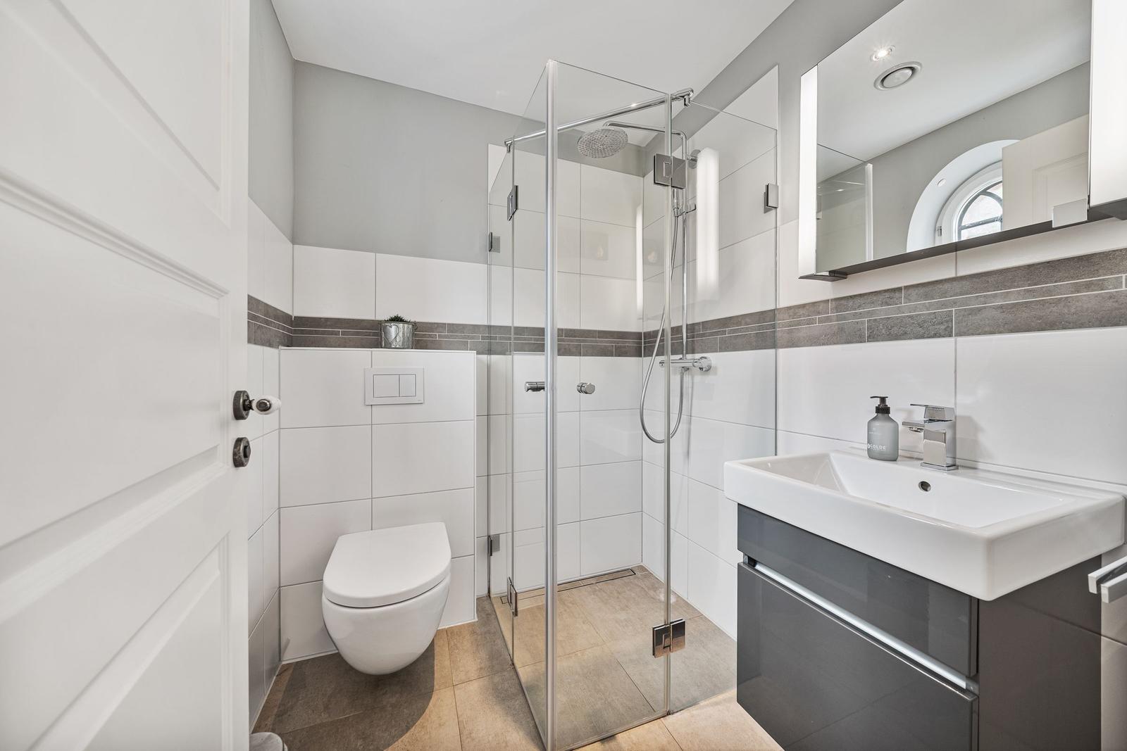 Badezimmer mit Dusche, Waschbecken und Toilette. Weiße Fliesen und graue Akzente.