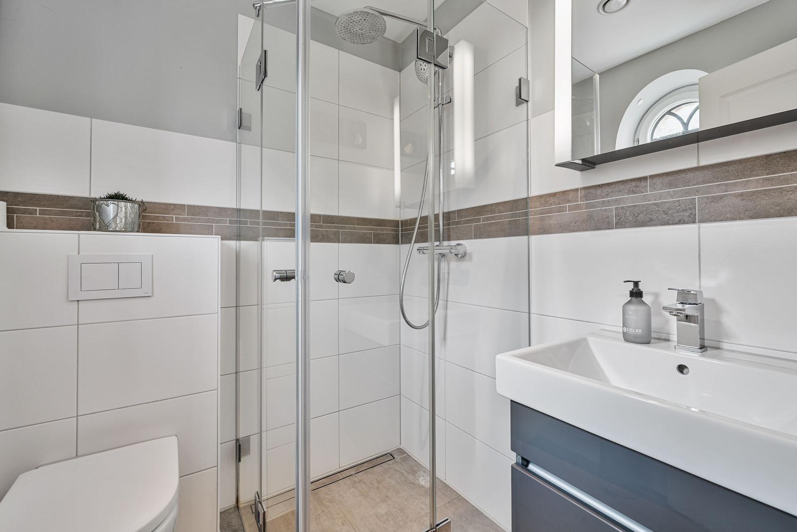 Badezimmer mit Dusche, Waschbecken und Toilette. Weiße Fliesen und graue Akzente.
