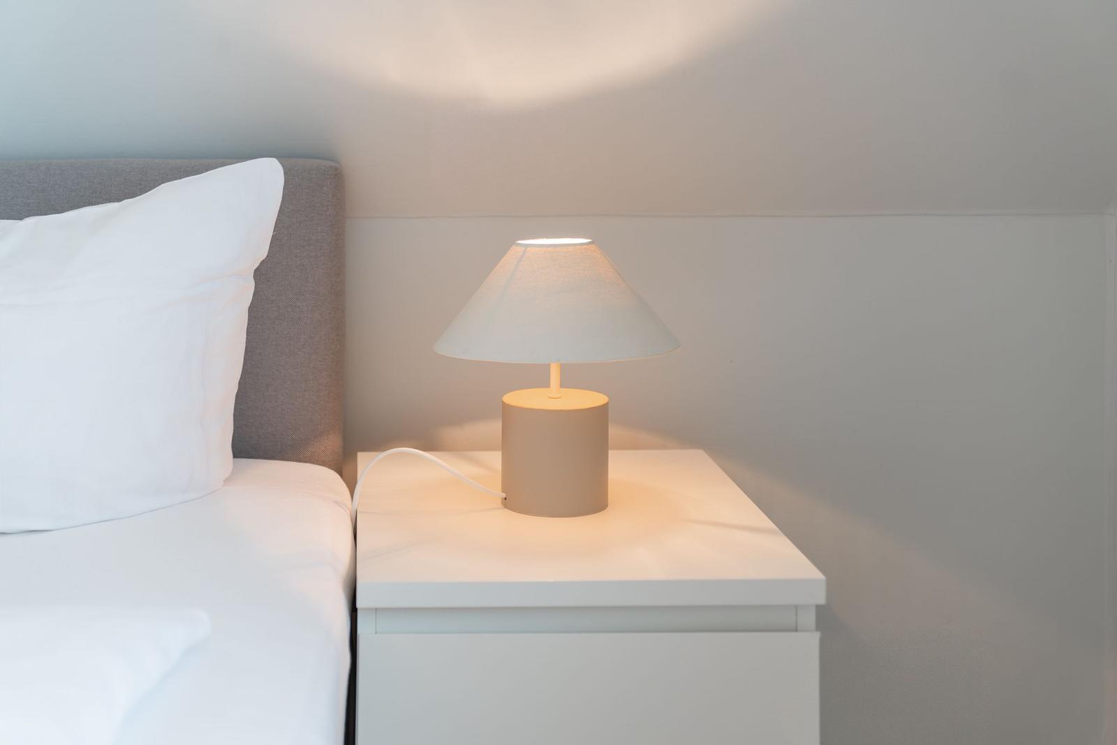 Nachttisch mit Lampe neben Bett mit Kissen und Bettwäsche.