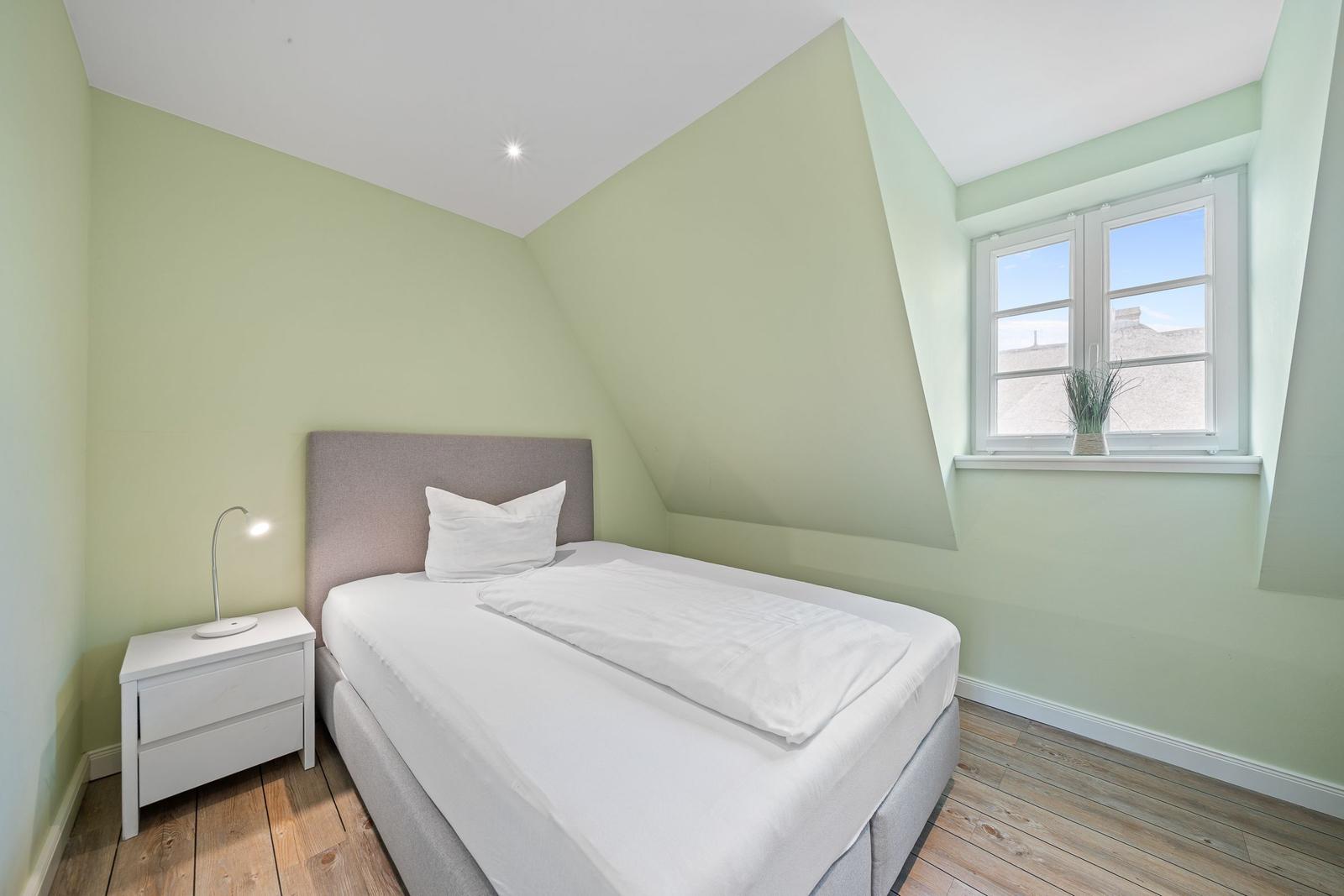 Schlafzimmer mit Dachschräge, weißem Bett und Nachttisch. Fenster mit Pflanze.