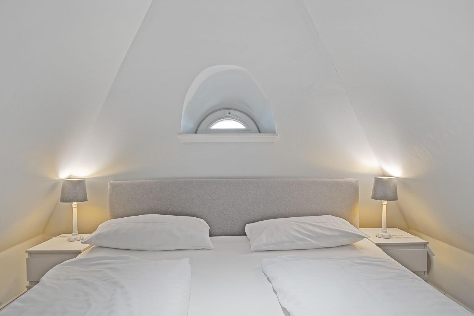 Doppelbett mit weißen Bettwäsche, zwei Nachttische und Lampe unter schrägen Dachfenstern.