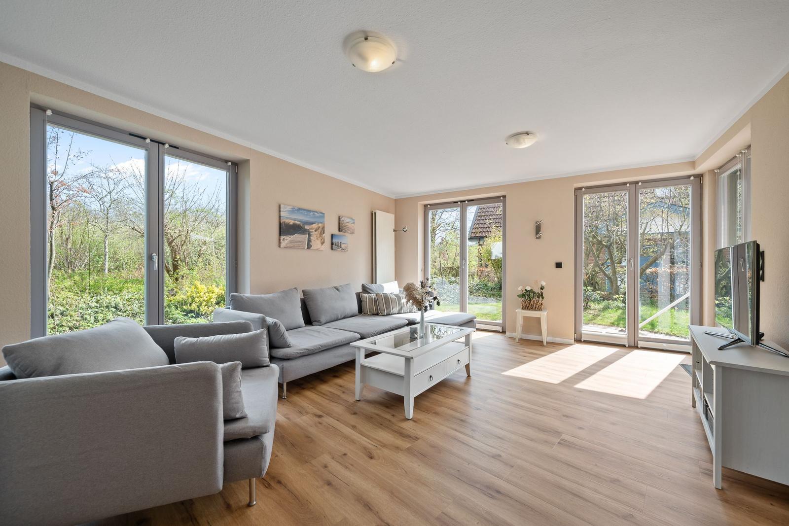 Wohnzimmer mit grauem Sofa, weißem Couchtisch und Holzboden. Große Fenster mit Blick auf Garten.
