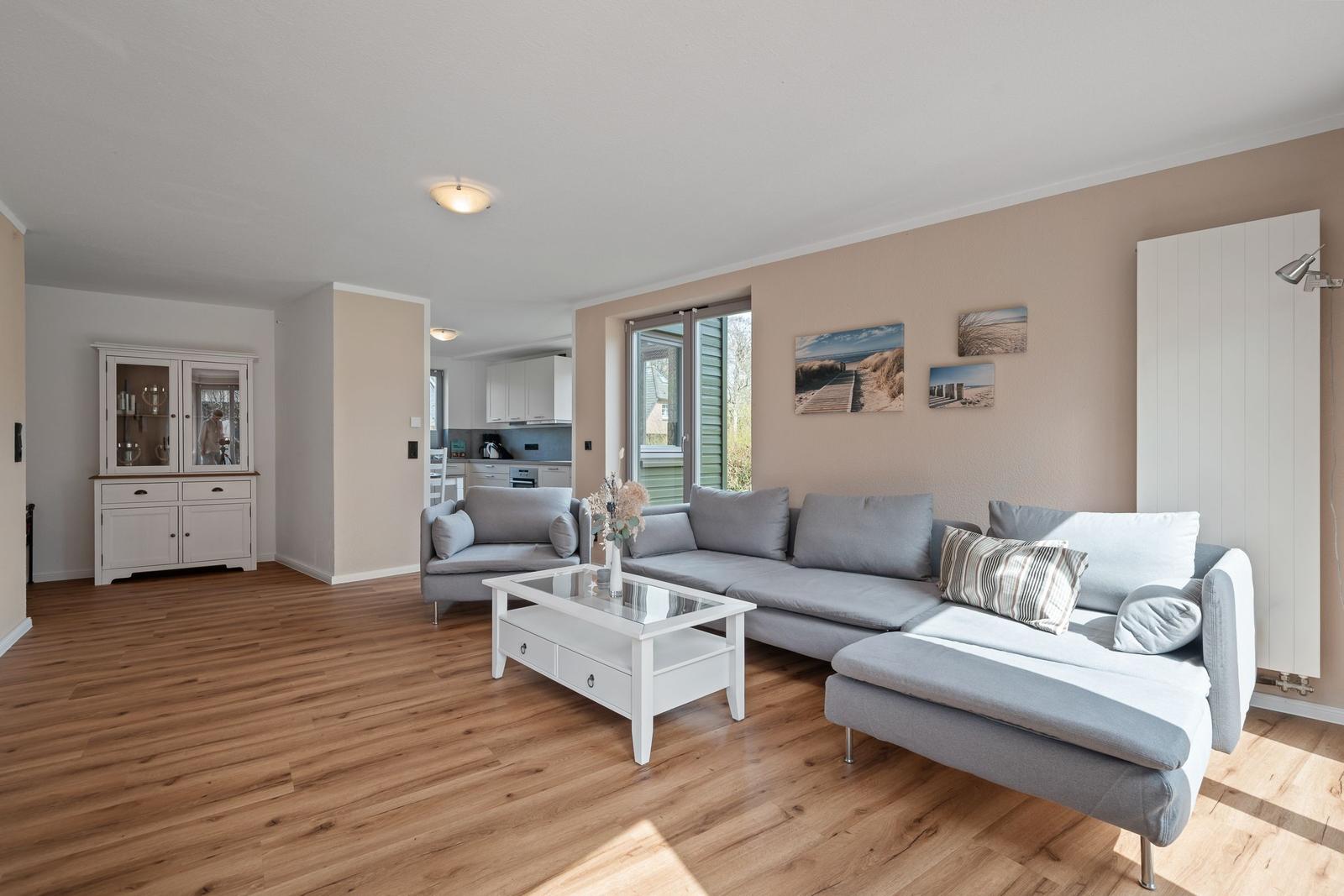 Wohnzimmer mit grauem Sofa, weißem Couchtisch und Holzboden. An der Wand hängen Strandbilder.
