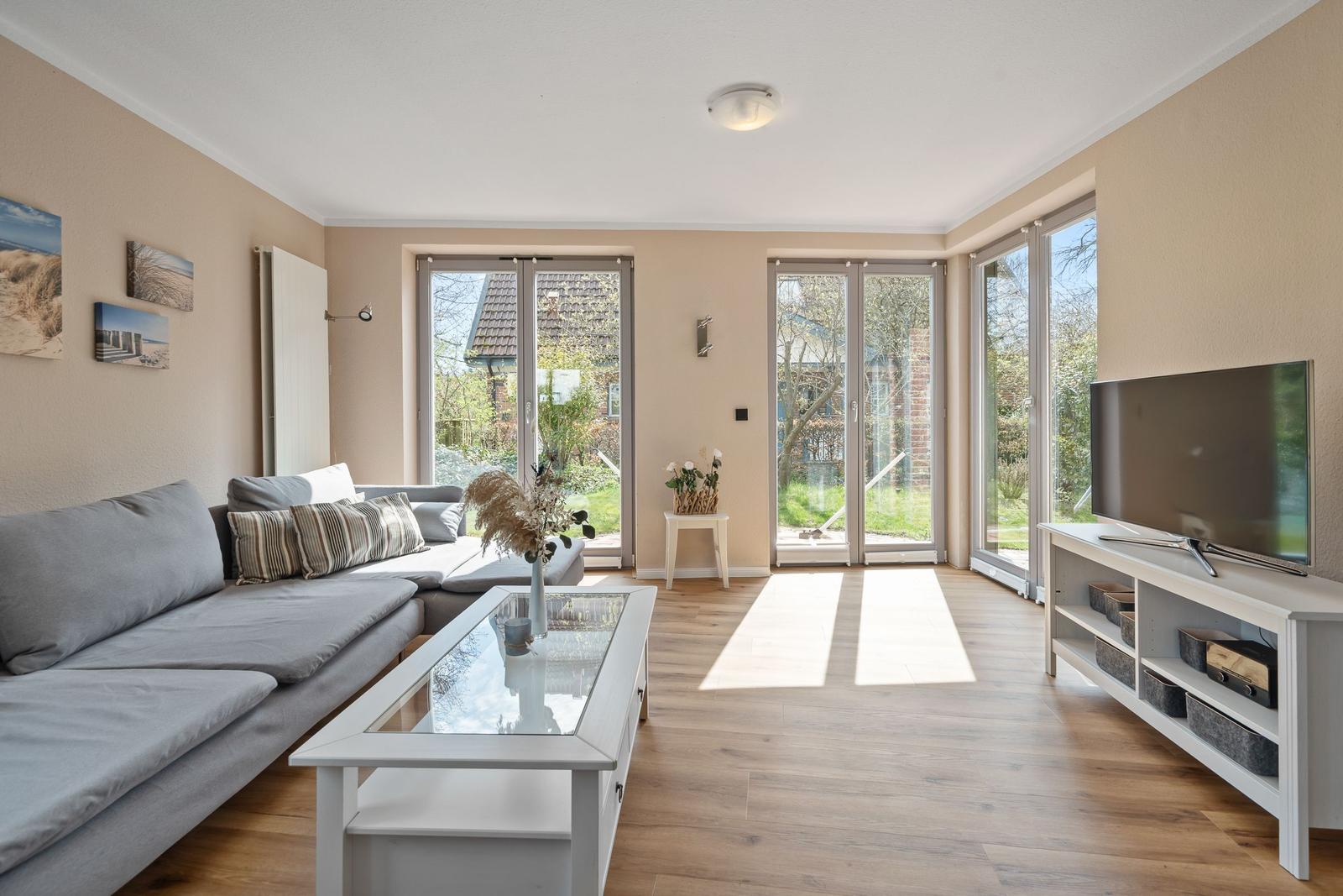 Wohnzimmer mit Sofa, Glas-Tisch und TV. Große Fenster mit Blick auf Garten.