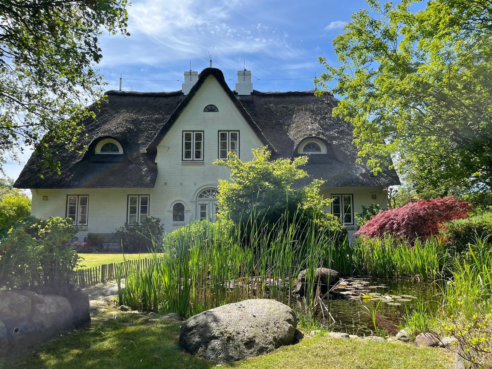 Weißes Haus mit Strohdach, umgeben von Garten und Teich.