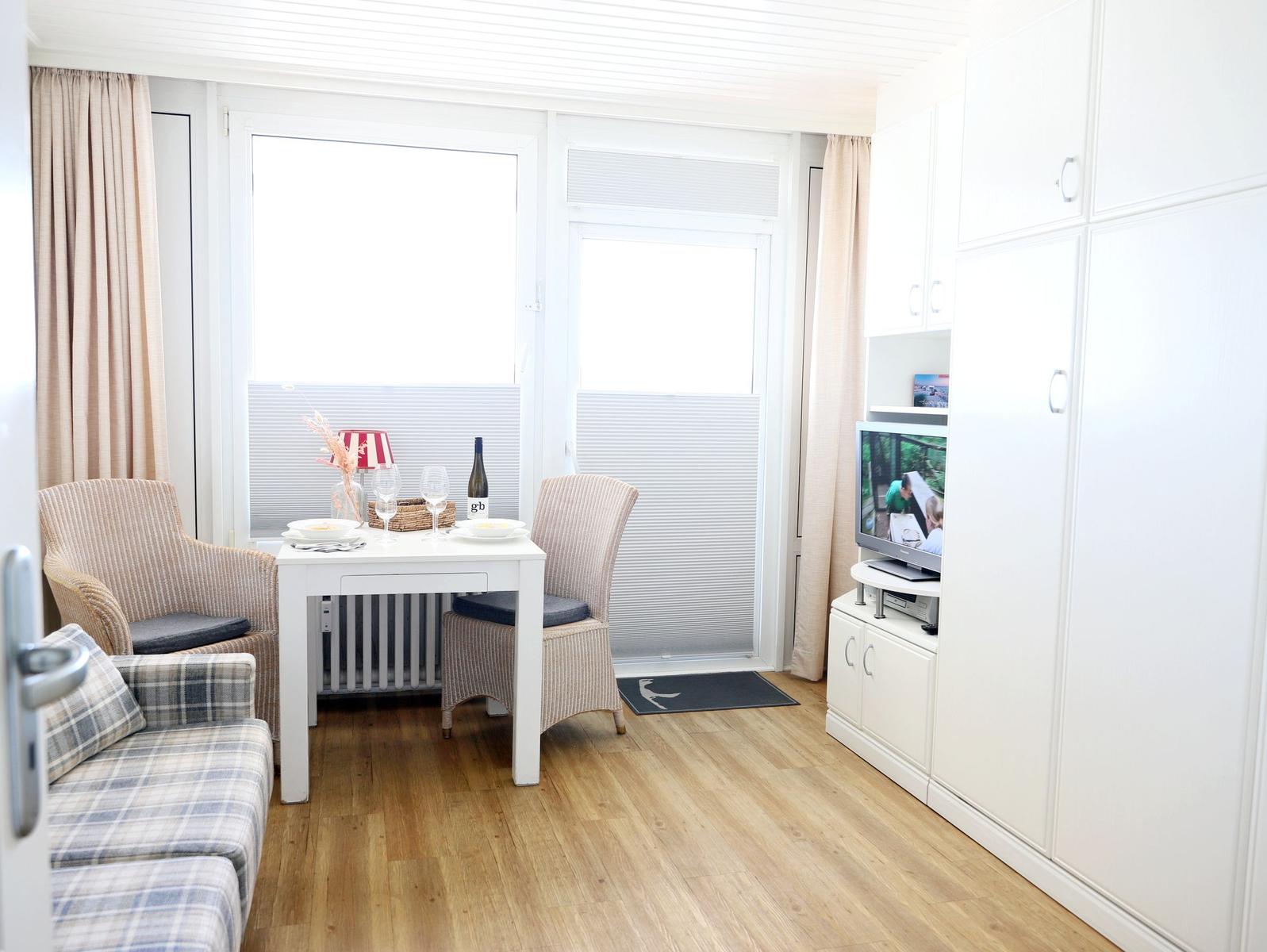Wohnraum mit Esstisch, zwei Stühlen, Sofa und Fernseher. Holzboden und große Fenster.
