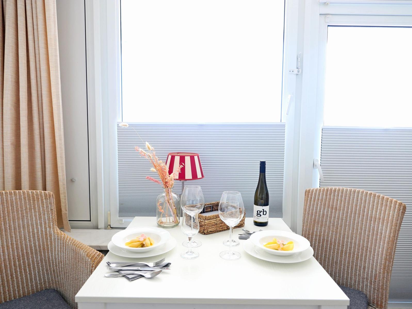 Tisch mit zwei Tellern, Gläsern und einer Weinflasche vor Fenster mit Jalousie.