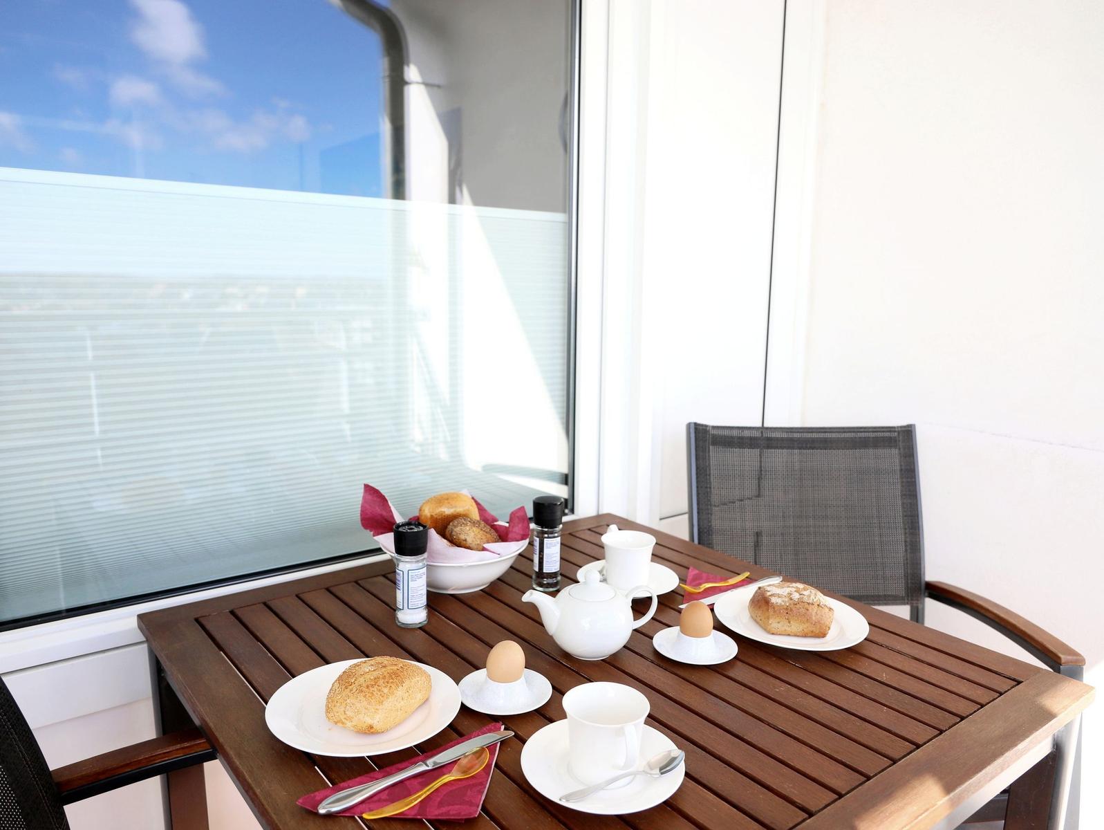 Terrasse mit Esstisch, Kaffeetasse, Brot und Eiern. Hintergrund: Glaswand und Himmel.