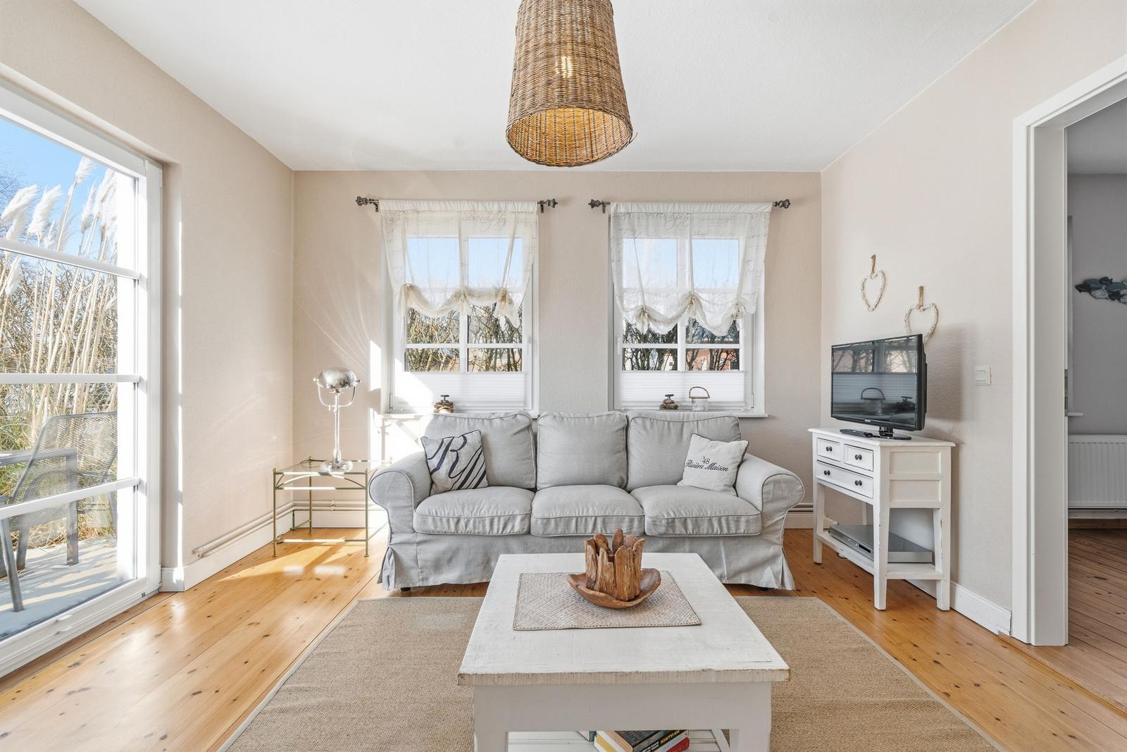 Wohnzimmer mit Sofa, Couchtisch und Fernseher. Große Fenster und Terrasse.