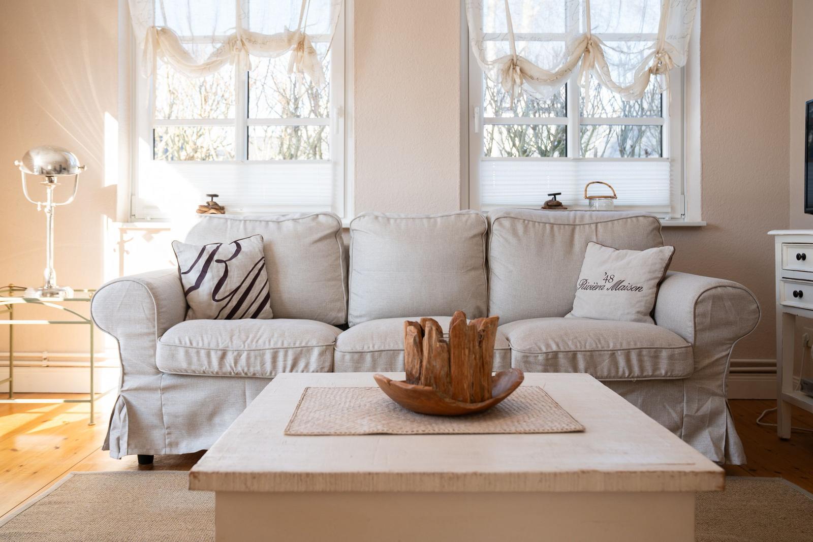 Ein Wohnzimmer mit einem hellen Sofa, zwei Fenstern und einem Holztisch mit einer Schale.