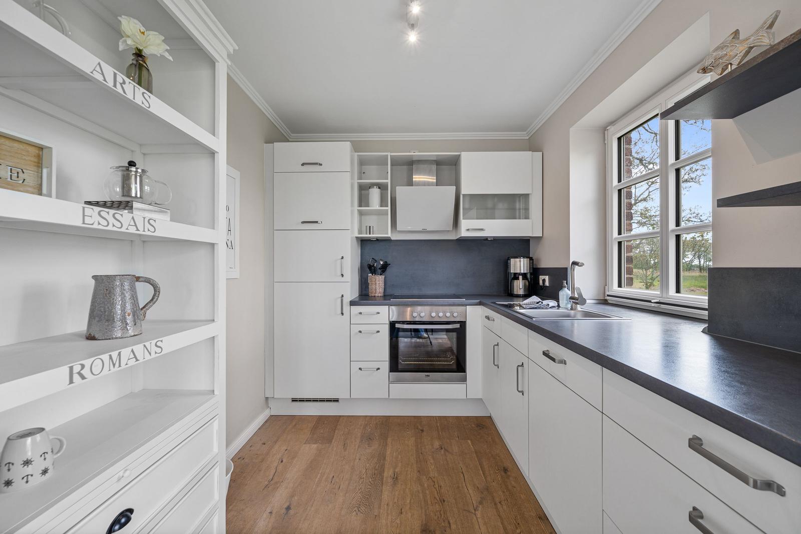 Moderne Küche mit weißen Schränken, dunkler Arbeitsplatte und Holzboden. Fenster mit Blick ins Freie.