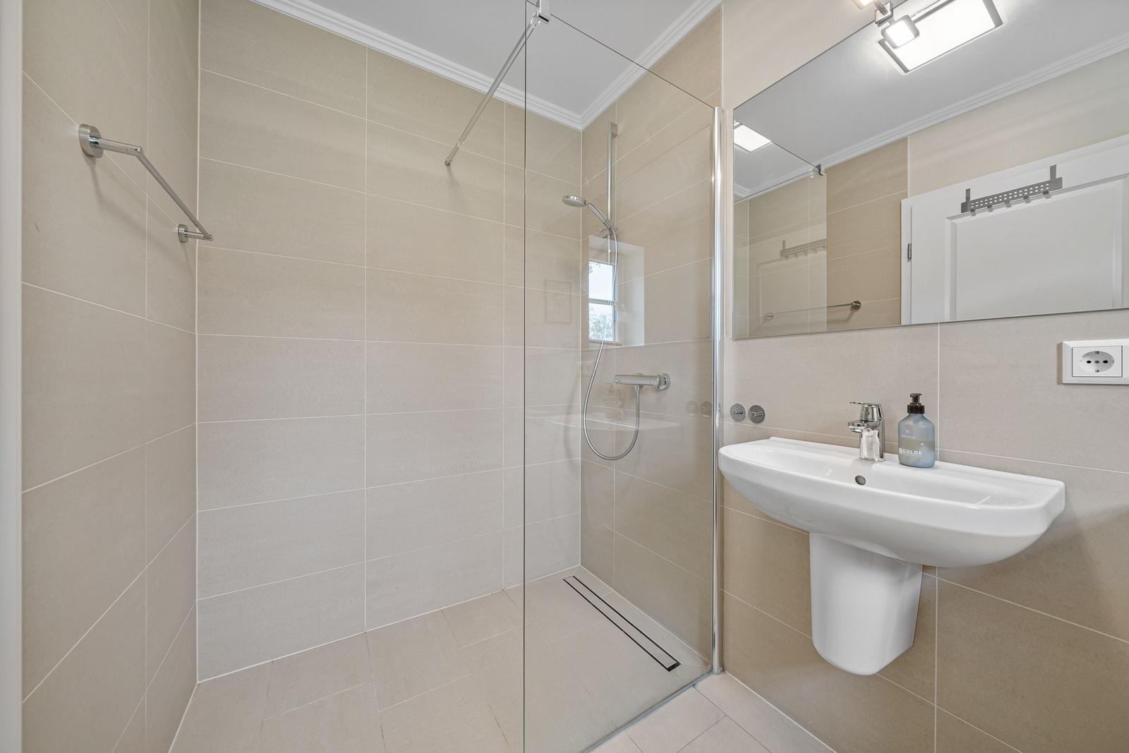 Dusche mit Glaswand, Waschbecken und Spiegel. Fliesenwand und Boden.