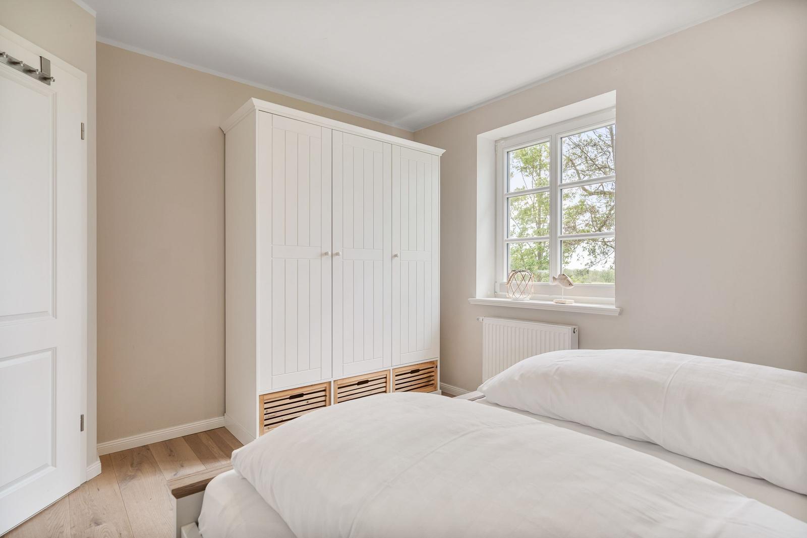 Schlafzimmer mit weißem Kleiderschrank, Bett und Fenster mit Blick auf Bäume.
