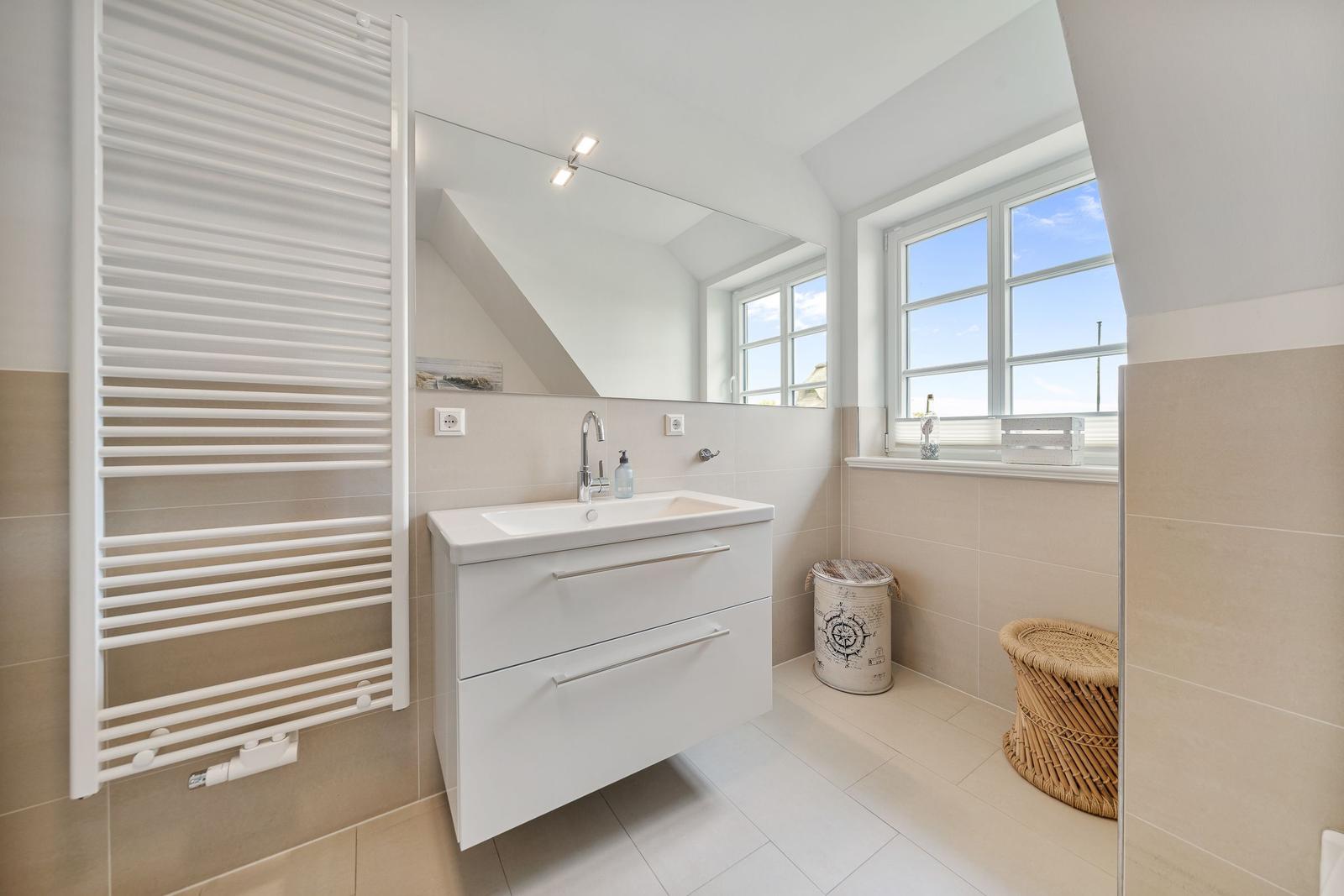 Badezimmer mit Waschbecken, Spiegel und Fenster. Weiße Einbauküche und Heizung.