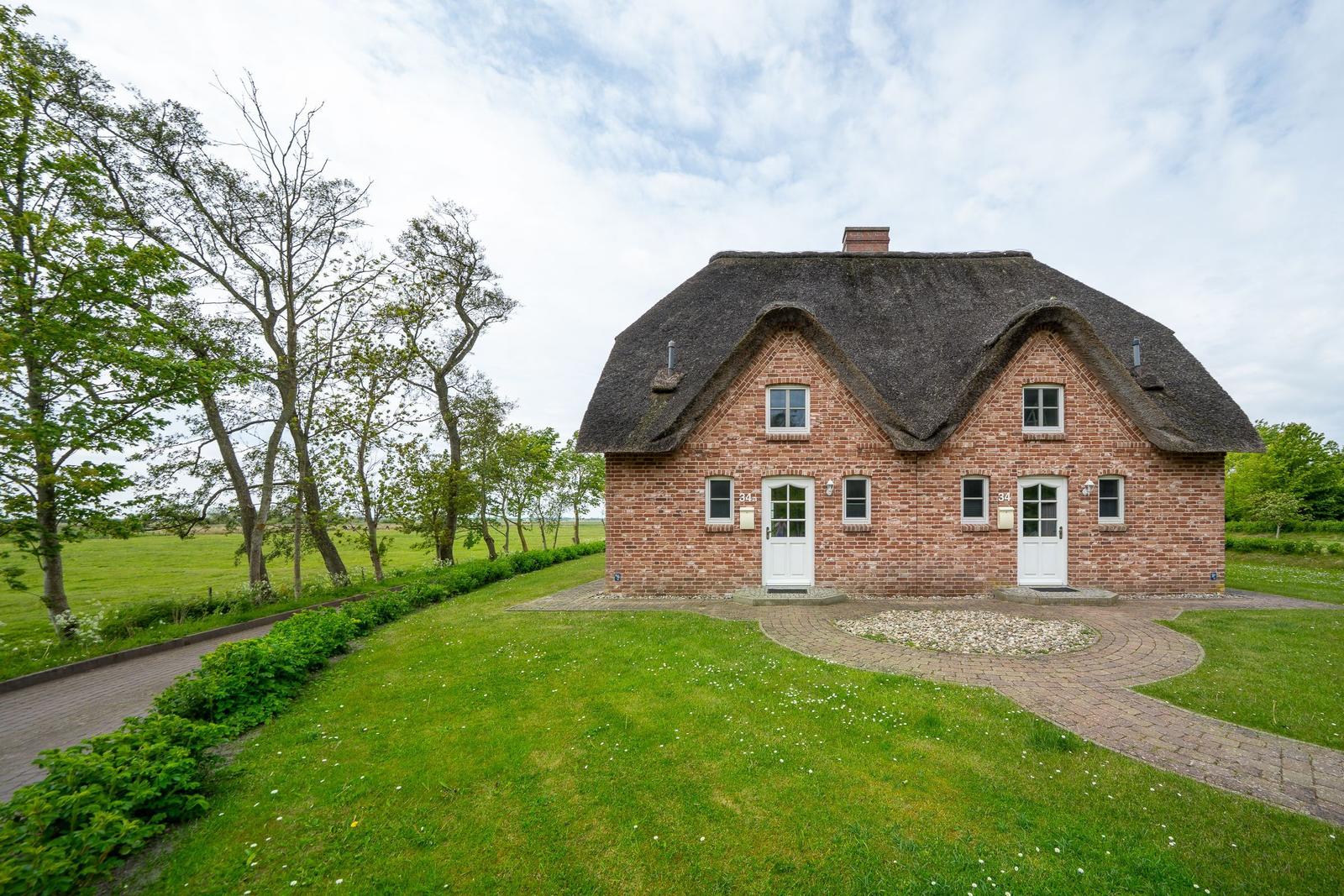 Rotes Backsteingebäude mit Strohdach und zwei Eingängen. Grünflächen und Bäume umgeben das Haus.