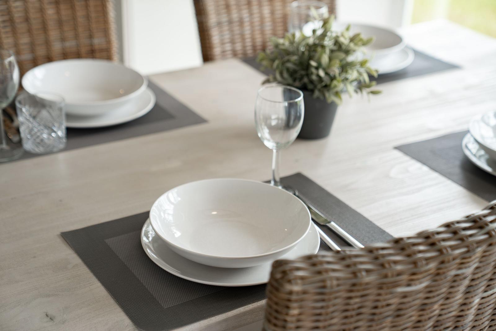 Tisch gedeckt mit weißen Tellern, Gläsern und dunklen Tischsets. Rattan-Stühle und Pflanze im Hintergrund.